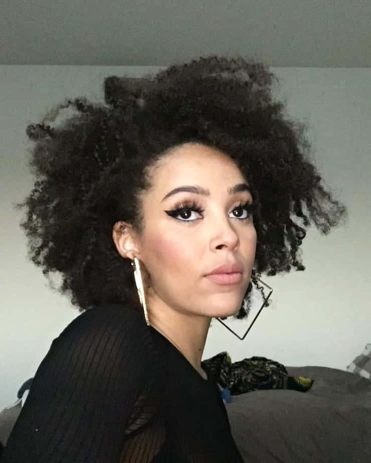 Doja Cat 桌布 壁紙 — 一位擁有豐盈深色捲髮和大膽翼狀眼線的女性在柔和光線的室內環境中直視鏡頭。