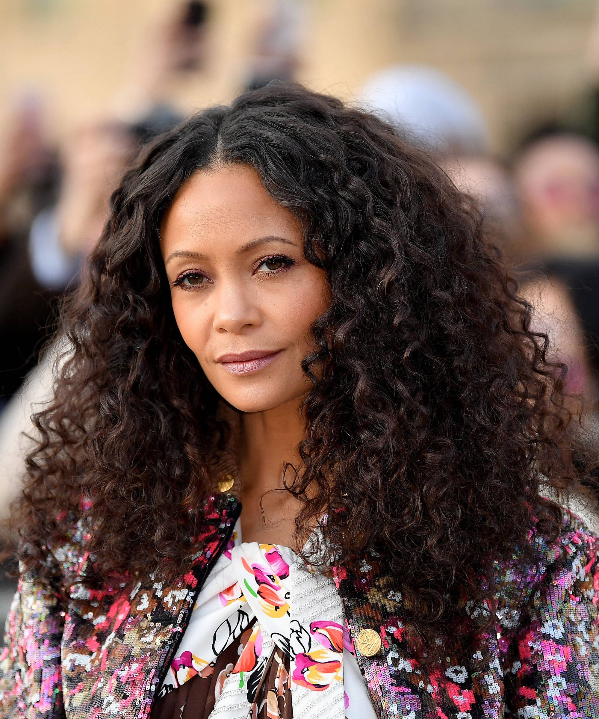 Actress HD 桌布 壁紙 — 一位擁有豐盈深色捲髮的女性，在模糊的人群背景下穿著色彩繽紛的亮片外套和花卉圖案上衣。