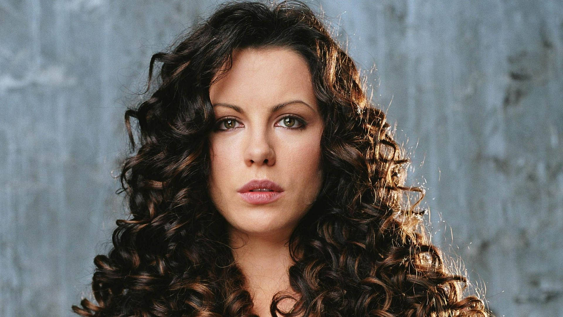 Kate Beckinsale 桌布 壁紙 — 一位擁有豐盈深色捲髮的女性，在紋理灰色牆壁前直視鏡頭。
