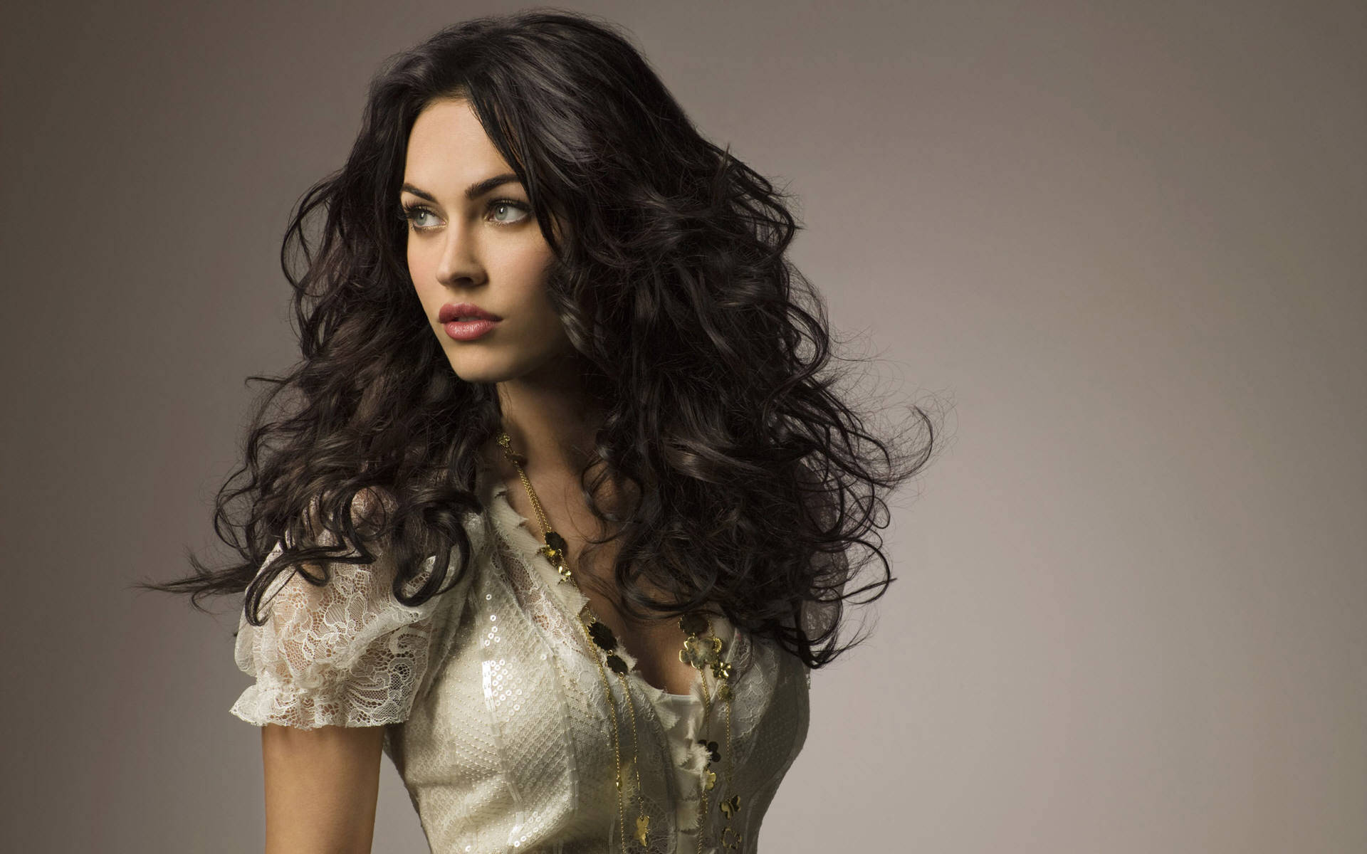 Megan Fox HD 桌布 壁紙 — 一位擁有豐盈深色捲髮的女性，穿著閃亮的奶油色蕾絲上衣，在工作室肖像中若有所思地凝視。