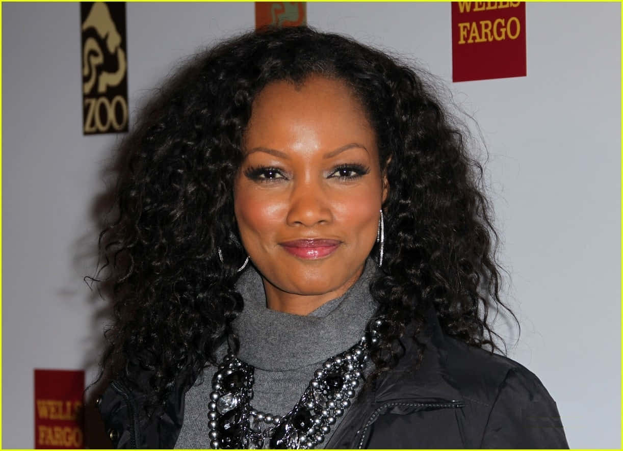 Garcelle Beauvais 桌布 壁紙 — 一位擁有豐盈深色捲髮的女性，穿著灰色高領毛衣和黑色外套，在紅毯活動上對著鏡頭溫暖微笑。