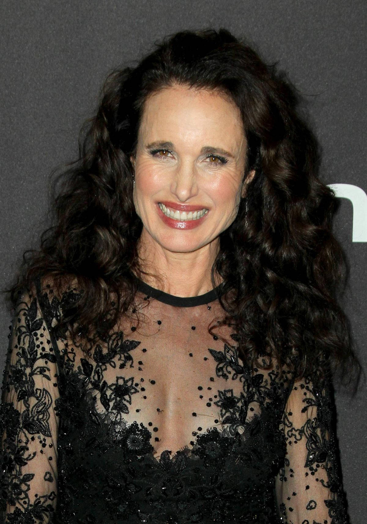andie macdowell 桌布 壁紙 — 一位擁有豐盈深色捲髮的耀眼女性，在正式活動中穿著黑色蕾絲禮服溫暖微笑。