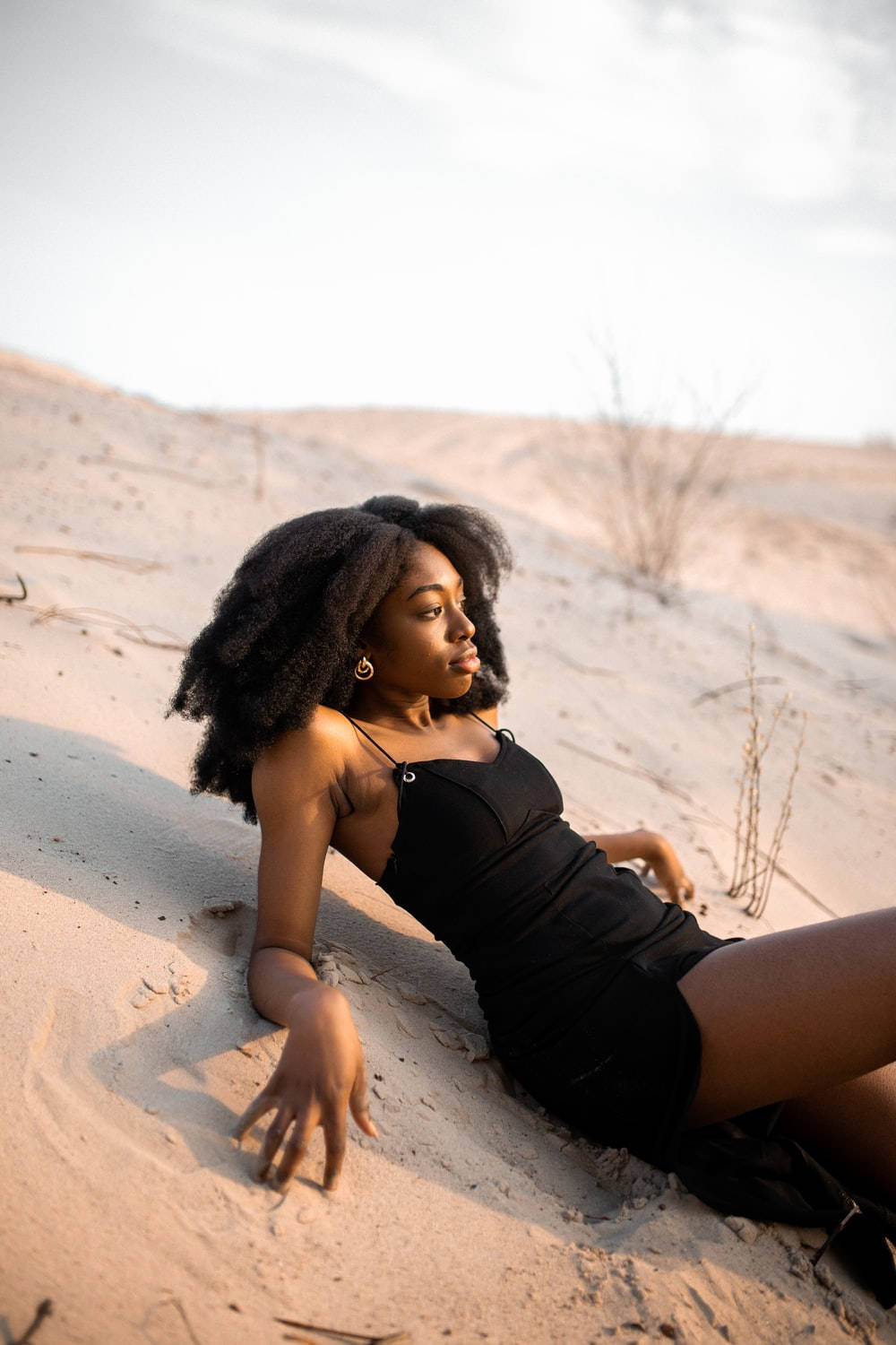Black Girl 桌布 壁紙 — 一位擁有豐盈深色頭髮的女性在柔和天空下優雅地躺在陽光照耀的沙丘上。
