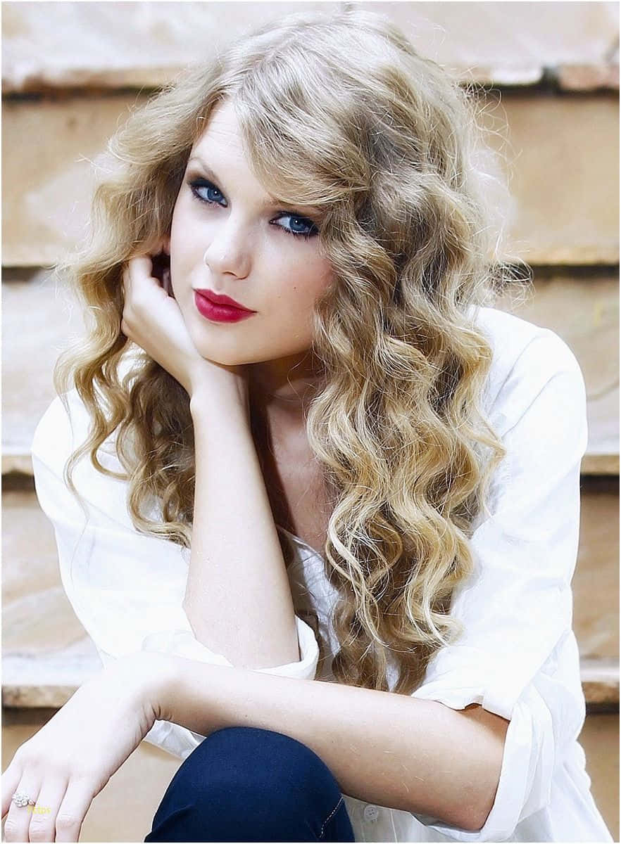 Taylor Swift iPhone 桌布 壁紙 — 一位擁有豐盈金色捲髮和大膽紅唇的女性，在木質背景下將下巴擱在手上。