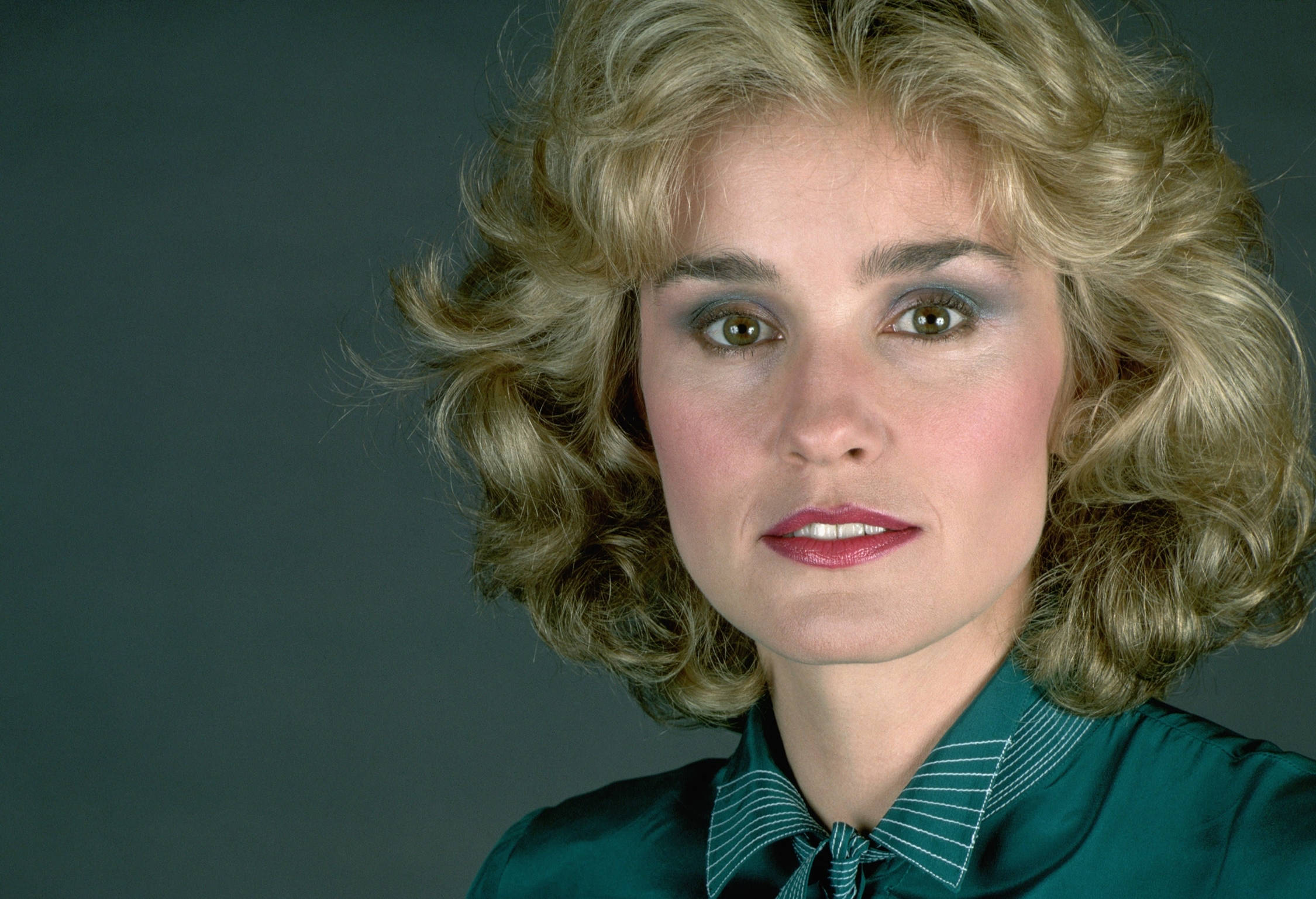 Jessica Lange 桌布 壁紙 — 一位擁有豐盈金色捲髮和鮮明妝容的女性，在柔和的灰色背景前直視鏡頭。