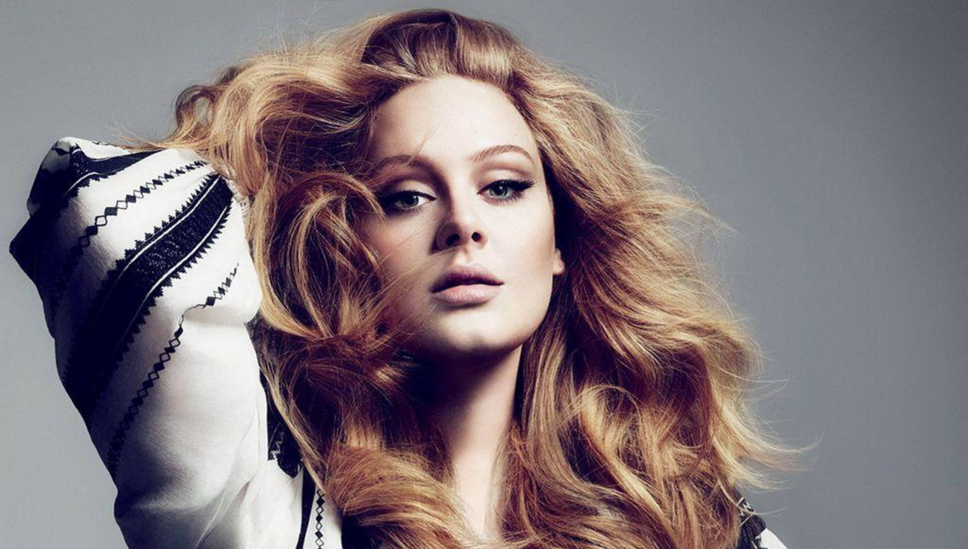 Adele 桌布 壁紙 — 一位擁有豐盈金色頭髮的女性，穿著黑白圖案袖子，在灰色背景下凝視著。