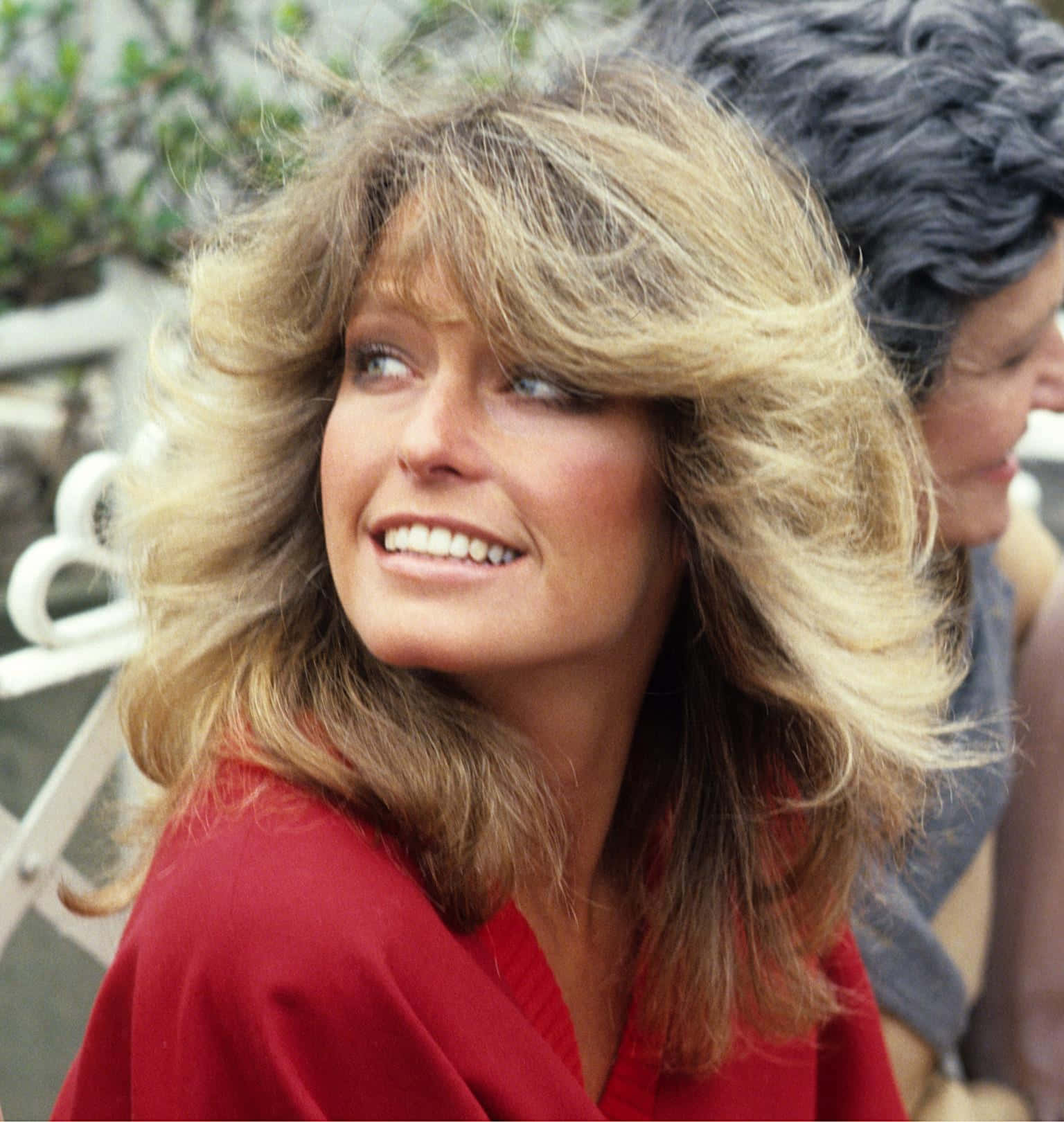 Farrah Fawcett 桌布 壁紙 — 一位擁有豐盈金髮和亮紅色上衣的女性，在戶外向上看時溫暖地微笑。