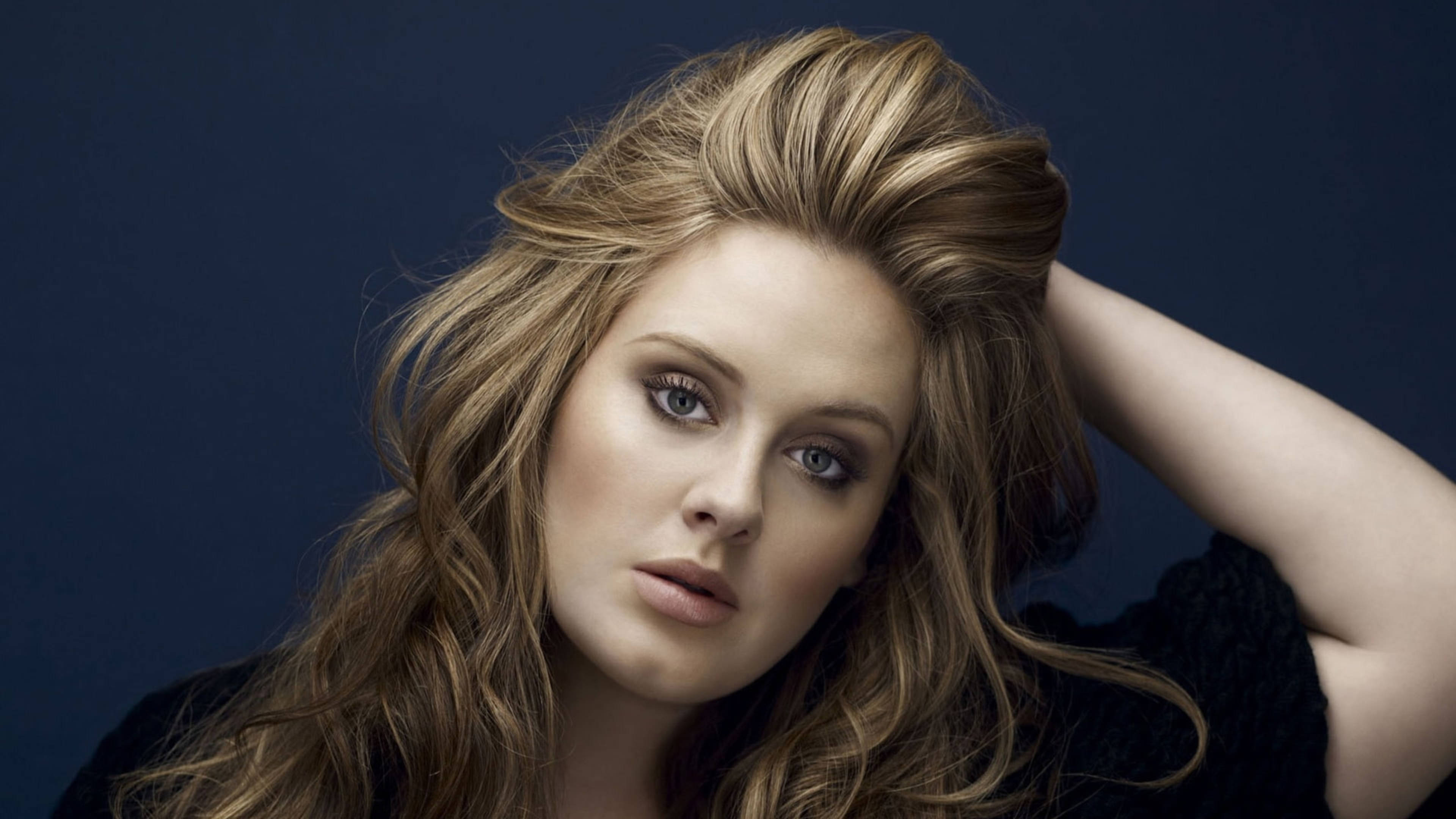 Adele 桌布 壁紙 — 一位擁有豐盈金髮和深邃藍眼睛的女性，在深藍色背景下直視鏡頭。