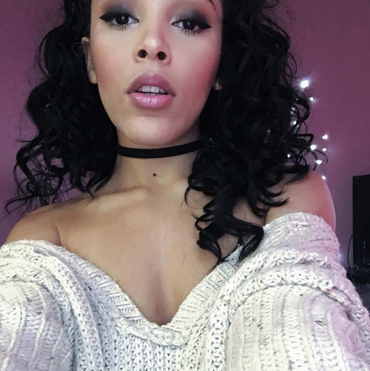 Doja Cat 桌布 壁紙 — 一位擁有豐盈黑色捲髮和粗眼線的女性，穿著一件厚實的白色針織毛衣，背景是柔和的粉紅色牆壁，上面裝飾著仙女燈。