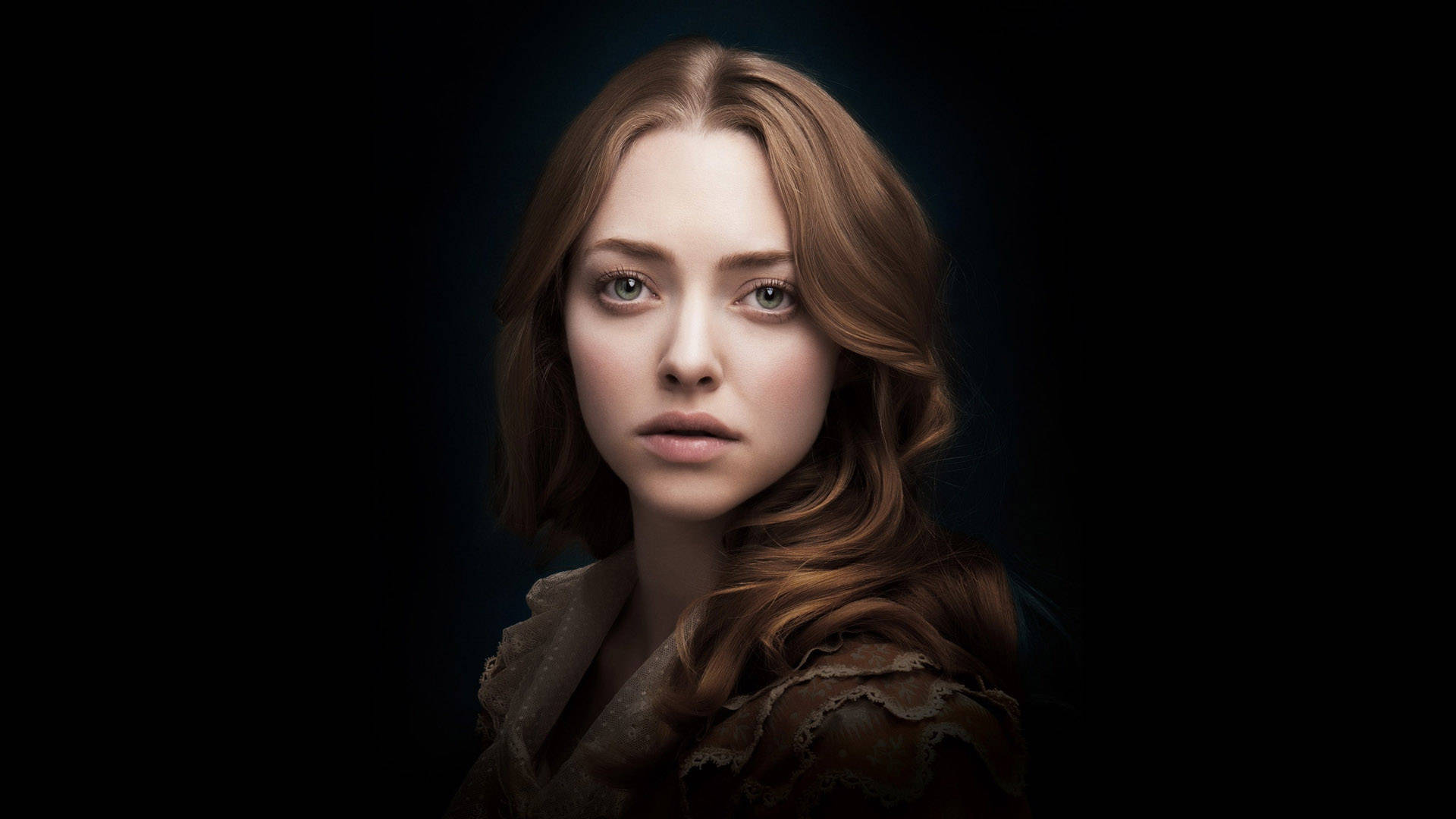 Amanda Seyfried 桌布 壁紙 — 一位擁有赤褐色頭髮和綠色眼睛的女性，在黑暗背景下直視前方，柔和的光線凸顯她的臉龐和質感服飾。