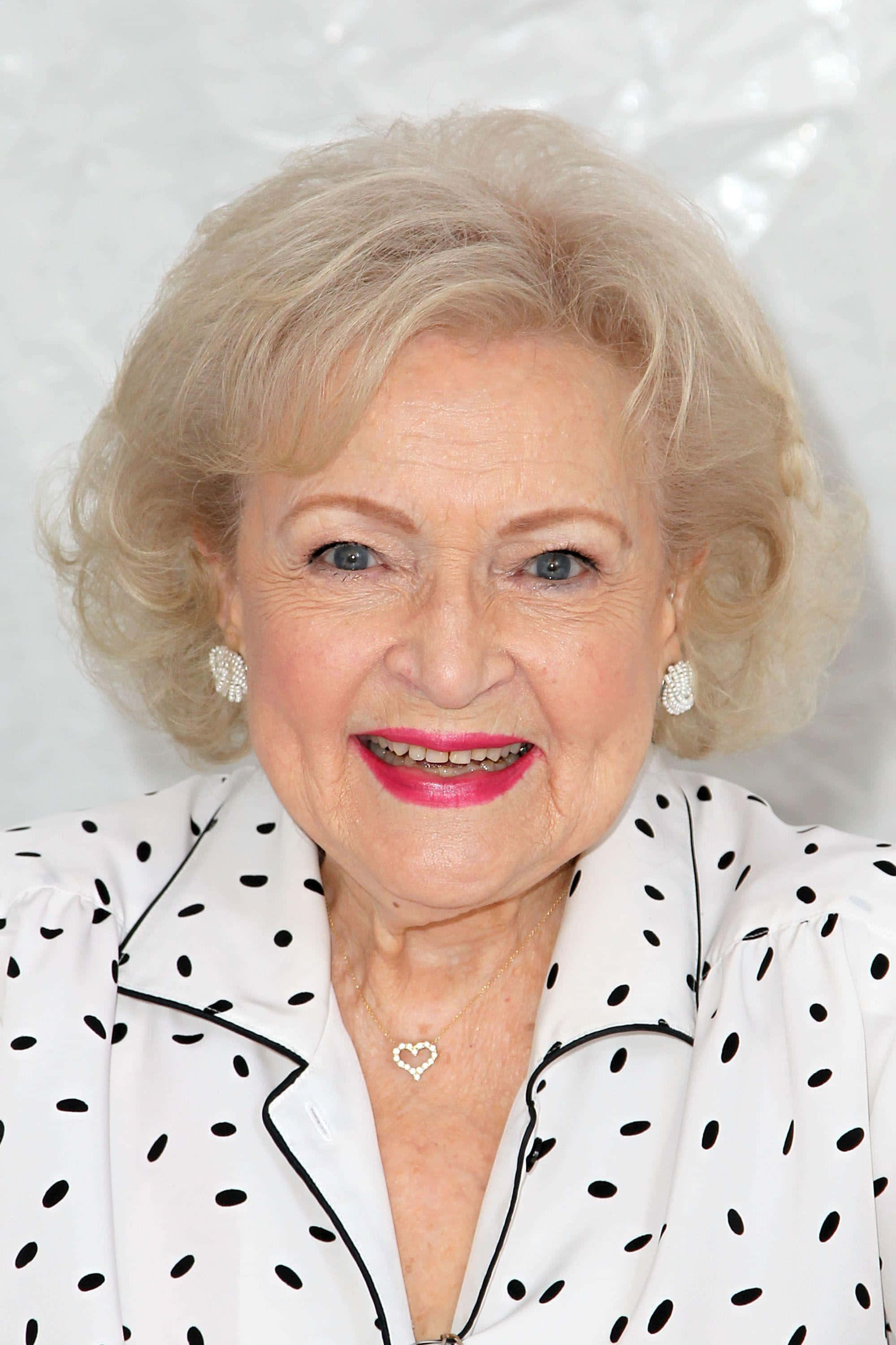 Betty White 桌布 壁紙 — 一位擁有金色捲髮和亮粉紅色唇膏的年長女性，穿著白色黑圓點襯衫，溫暖地微笑。