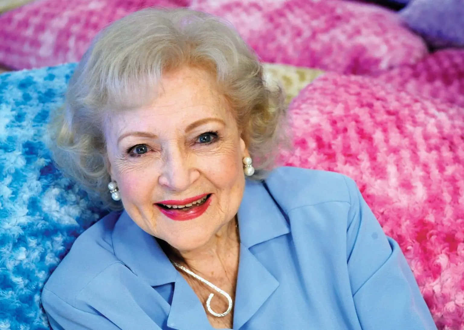 Betty White 桌布 壁紙 — 一位擁有金色捲髮和紅色唇膏的老年女性，靠在色彩繽紛、有紋理的枕頭上，露出溫暖的笑容。