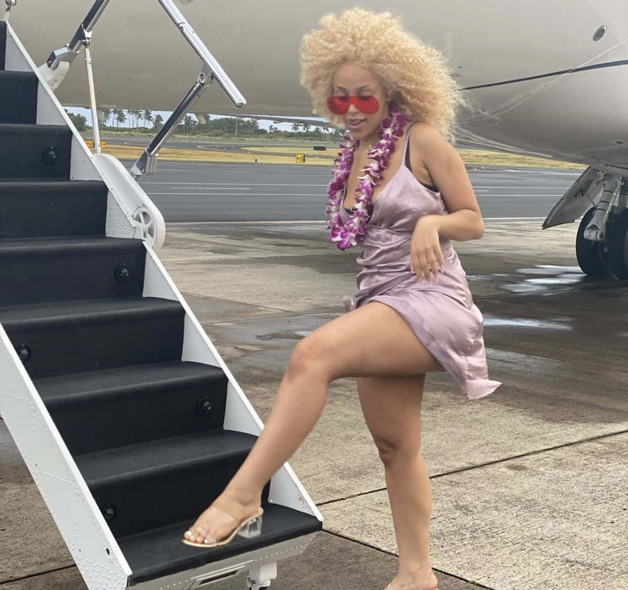 Doja Cat 桌布 壁紙 — 一位擁有金色捲髮和紅色太陽眼鏡的女性，穿著紫色連衣裙和花環走下飛機樓梯。