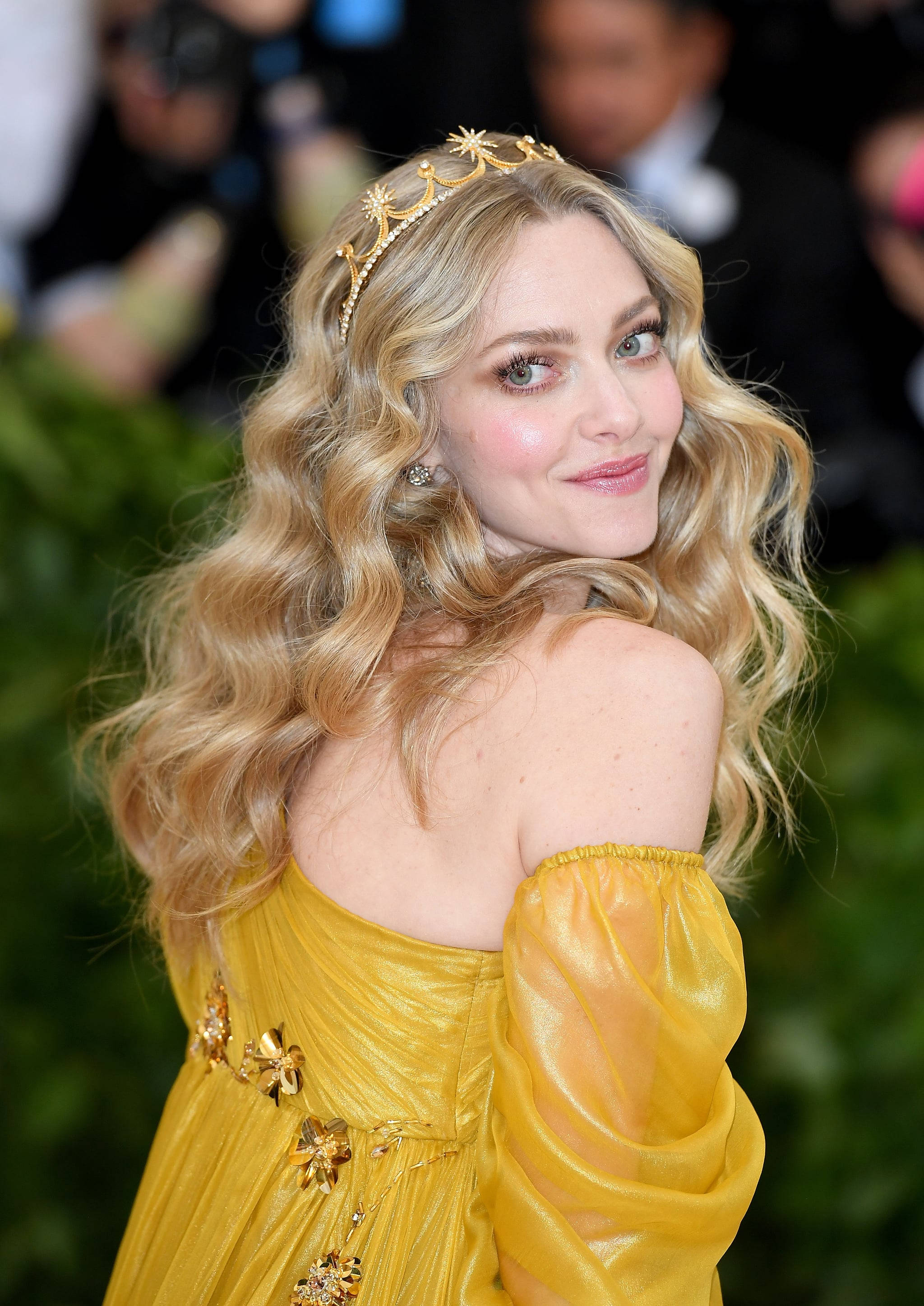 Amanda Seyfried 桌布 壁紙 — 一位擁有金色捲髮和鑲有寶石皇冠的女性，穿著閃閃發光的黃色禮服，在華麗的活動中回頭微笑。