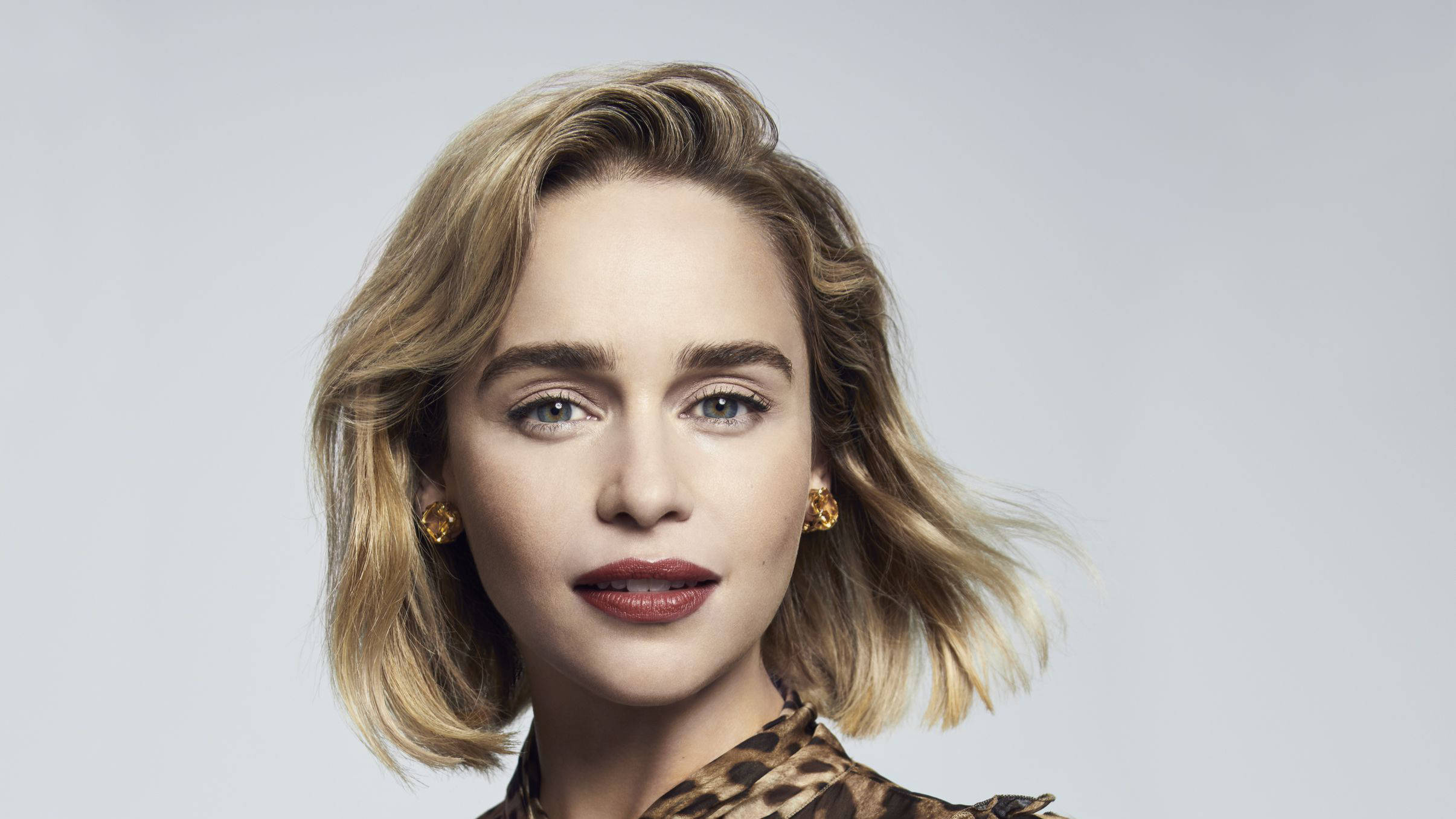 Emilia 桌布 壁紙 — 一位擁有金色波浪短髮和大膽紅唇的女性，在柔和的灰色工作室背景前直視鏡頭。