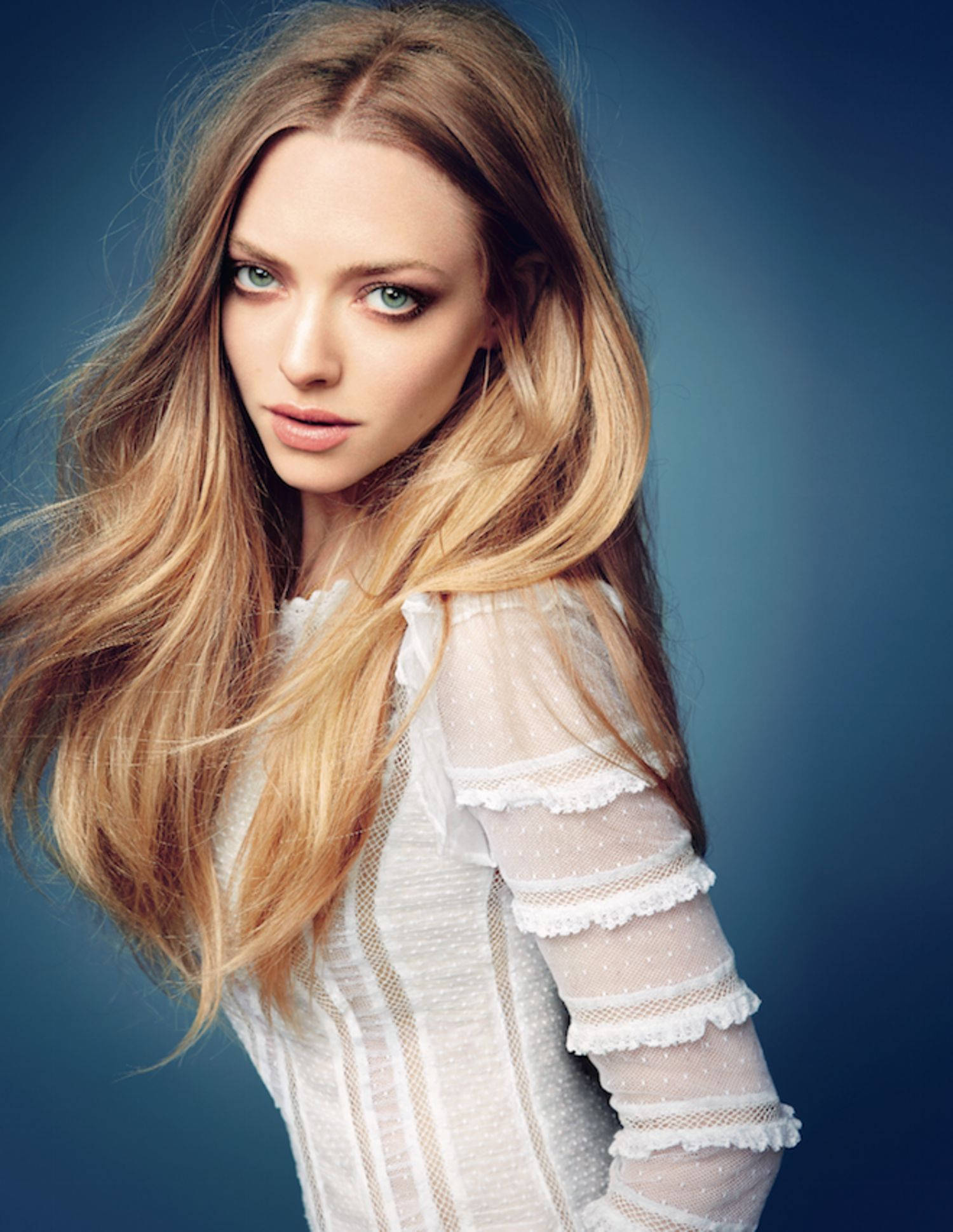Amanda Seyfried 桌布 壁紙 — 一位擁有金色長髮和引人注目的綠色眼睛的女性，身穿白色蕾絲邊上衣，在深藍色攝影棚背景下直視鏡頭。