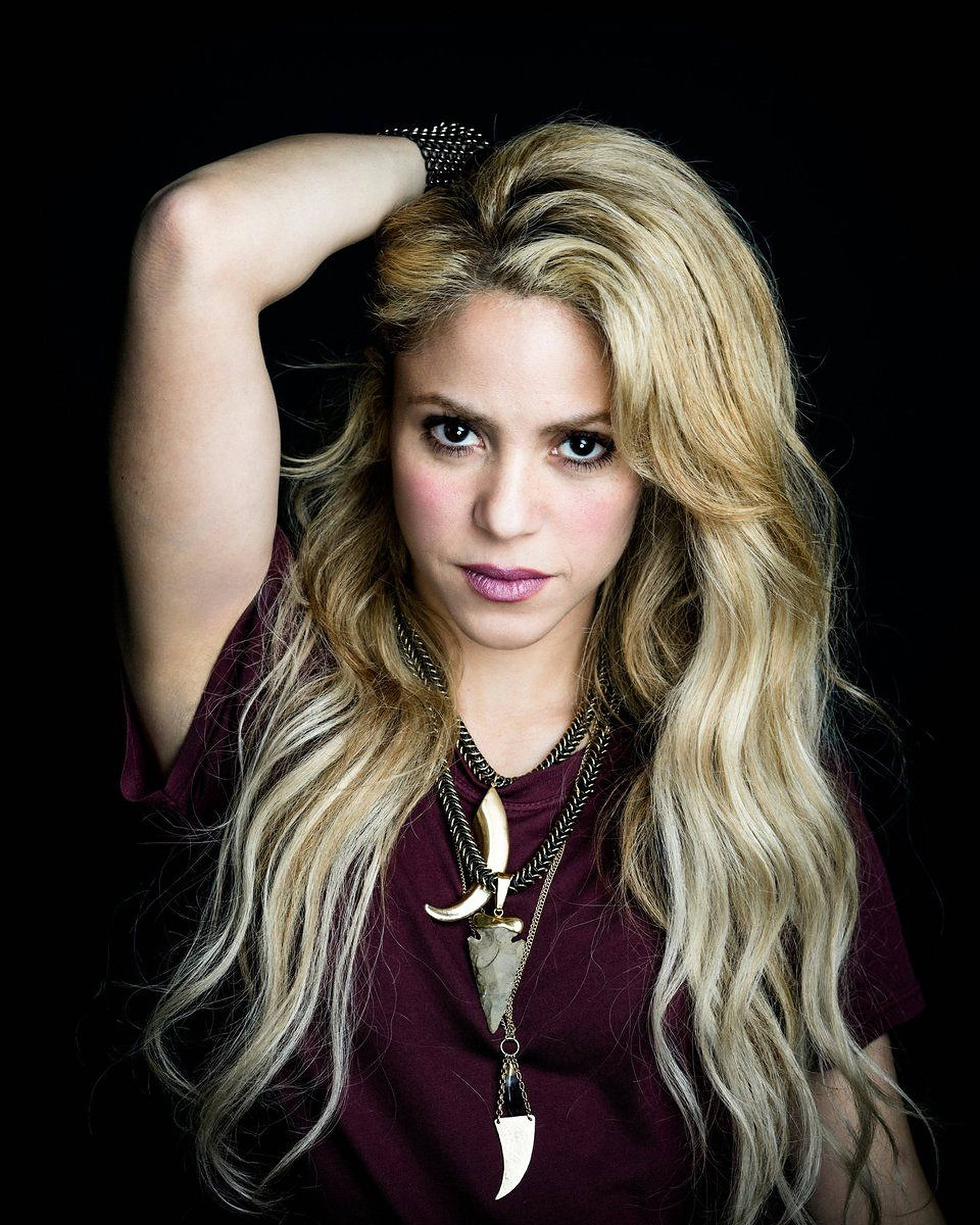 Shakira 桌布 壁紙 — 一位擁有金色長髮和濃妝的女性，在黑色背景前擺姿勢，穿著栗色襯衫和部落風格項鍊。