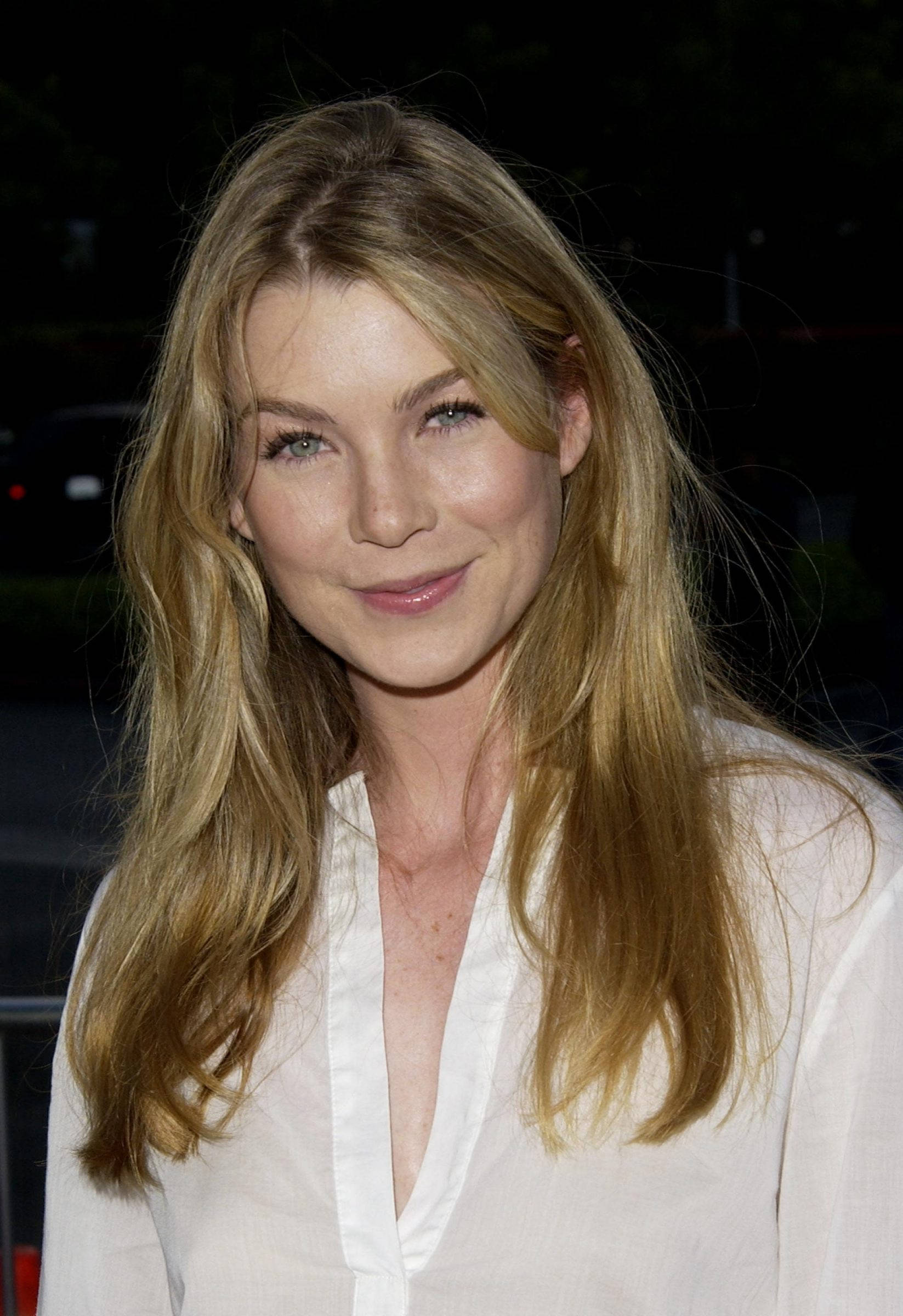 Ellen Pompeo 桌布 壁紙 — 一位擁有金色長髮和綠色眼睛的女性，在黑暗的夜間背景下穿著白色襯衫，溫柔地微笑。