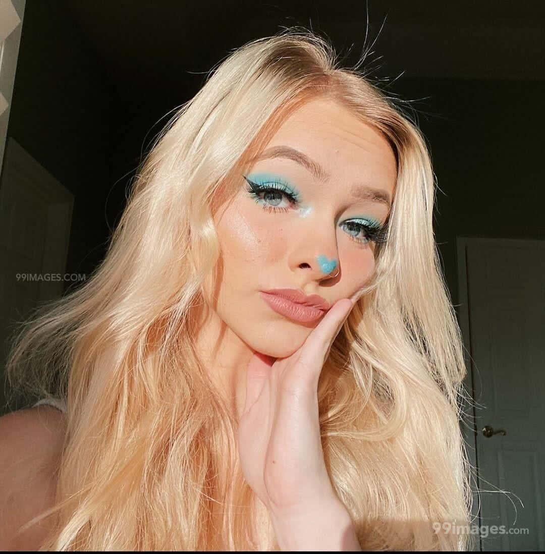 Zoe Laverne 桌布 壁紙 — 一位擁有金色長髮和鮮豔藍色眼妝的女性，在昏暗的房間裡將手放在下巴下擺姿勢。