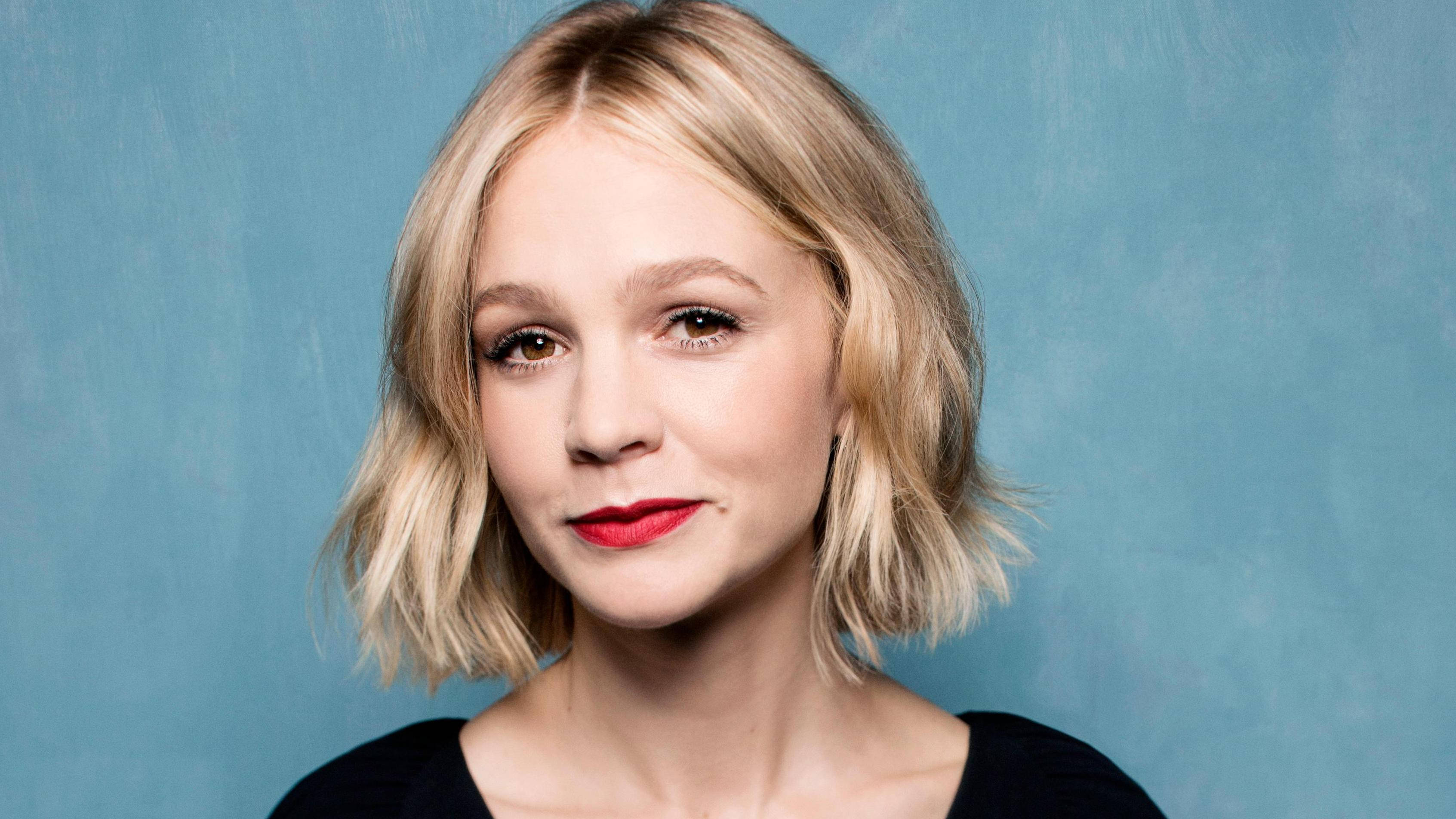Carey Mulligan 桌布 壁紙 — 一位擁有金色鮑伯頭和鮮豔紅唇的女性，在帶有紋理的藍色背景前溫柔凝視。