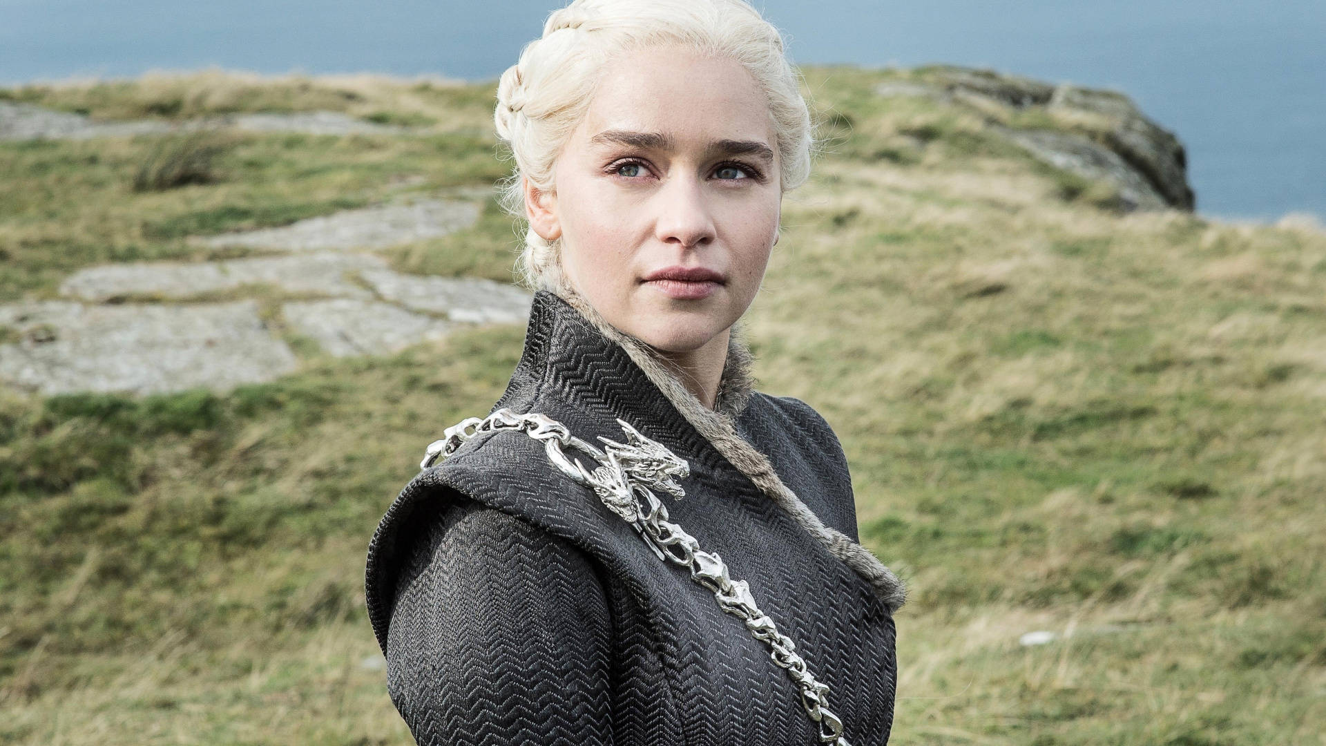 Emilia 桌布 壁紙 — 一位擁有鉑金色辮子頭髮和龍鏈的女人站在俯瞰大海的草崖上。