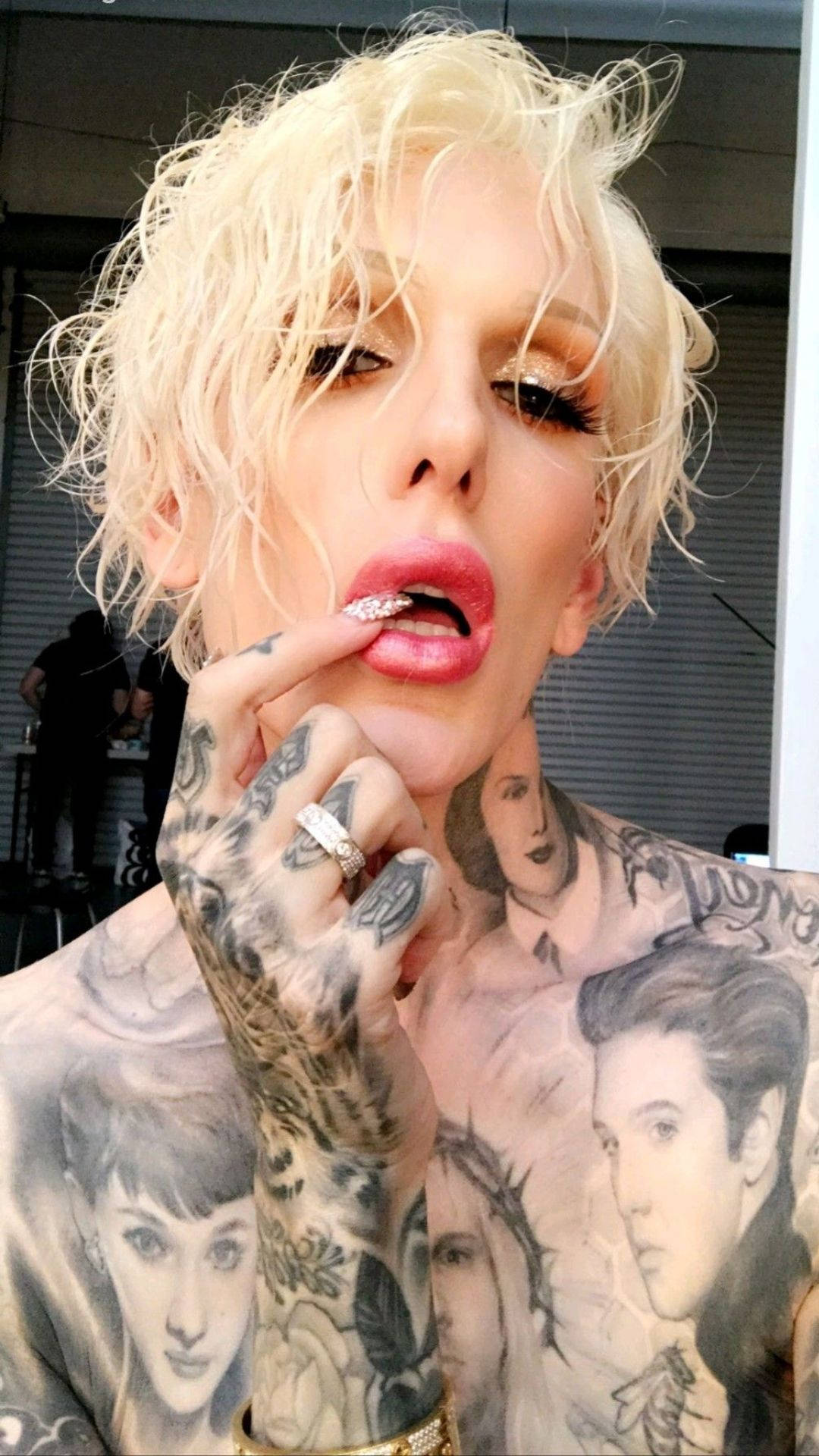 Jeffree Star 桌布 壁紙 — 一位擁有鉑金色頭髮、身上和手上紋有面孔紋身的人，用閃亮的指甲觸碰嘴唇。