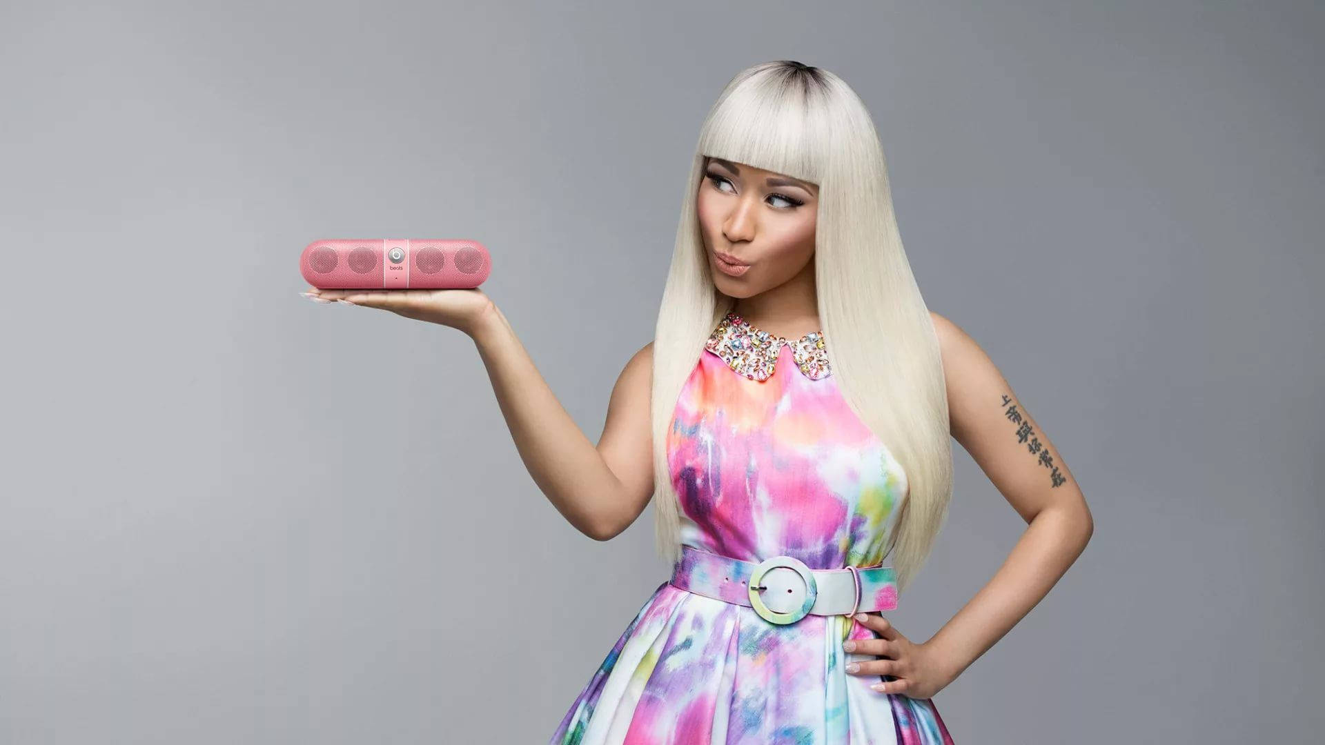 Nicki 桌布 壁紙 — 一位擁有鉑金色頭髮、身穿彩色連衣裙的女性，在純灰色背景前拿著一個粉紅色便攜式揚聲器。