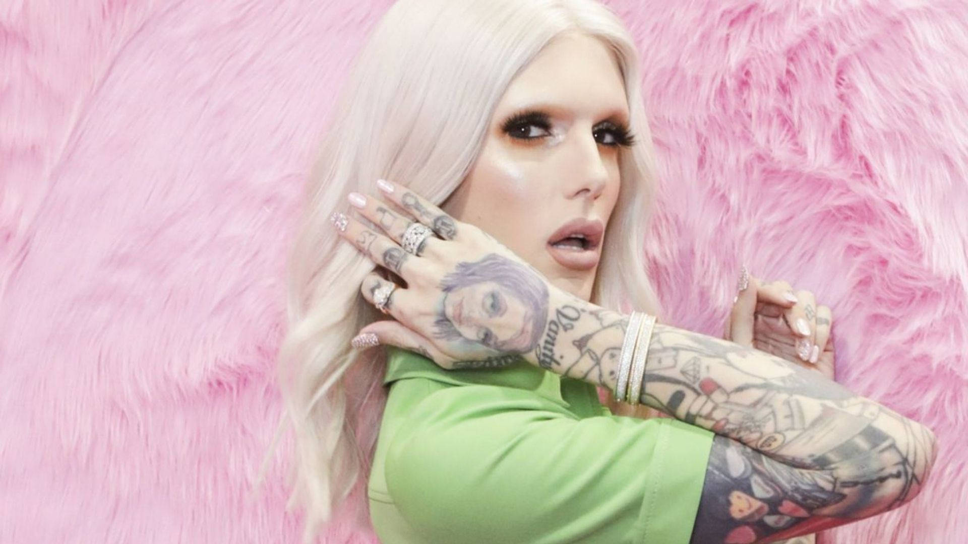 Jeffree Star 桌布 壁紙 — 一位擁有鉑金色頭髮和刺青的人，在毛絨粉紅色背景前穿著綠色上衣擺姿勢。