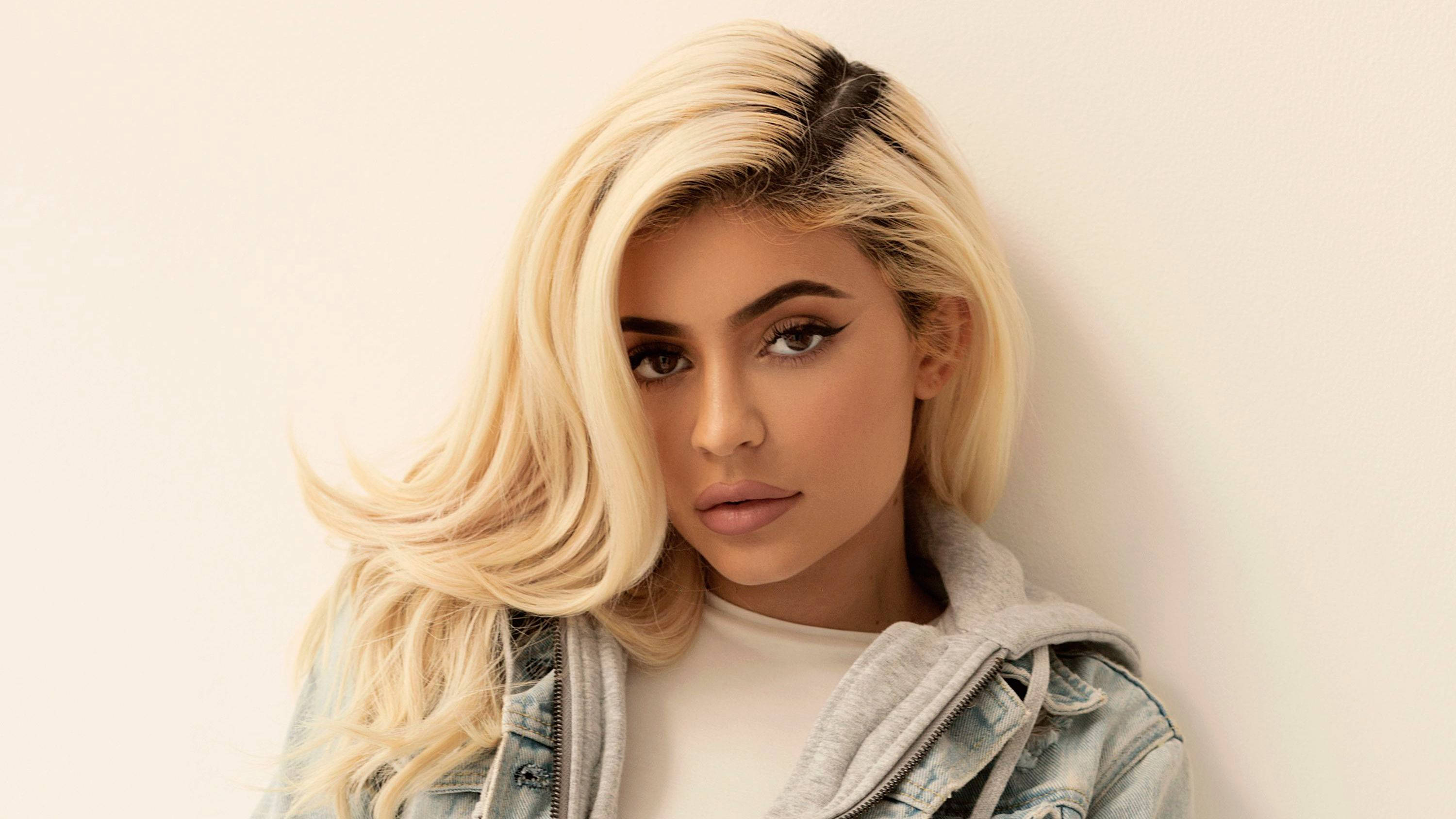 Kylie 桌布 壁紙 — 一位擁有鉑金色頭髮和大膽妝容的女性靠在一面素牆上，身穿牛仔外套內搭連帽衫。