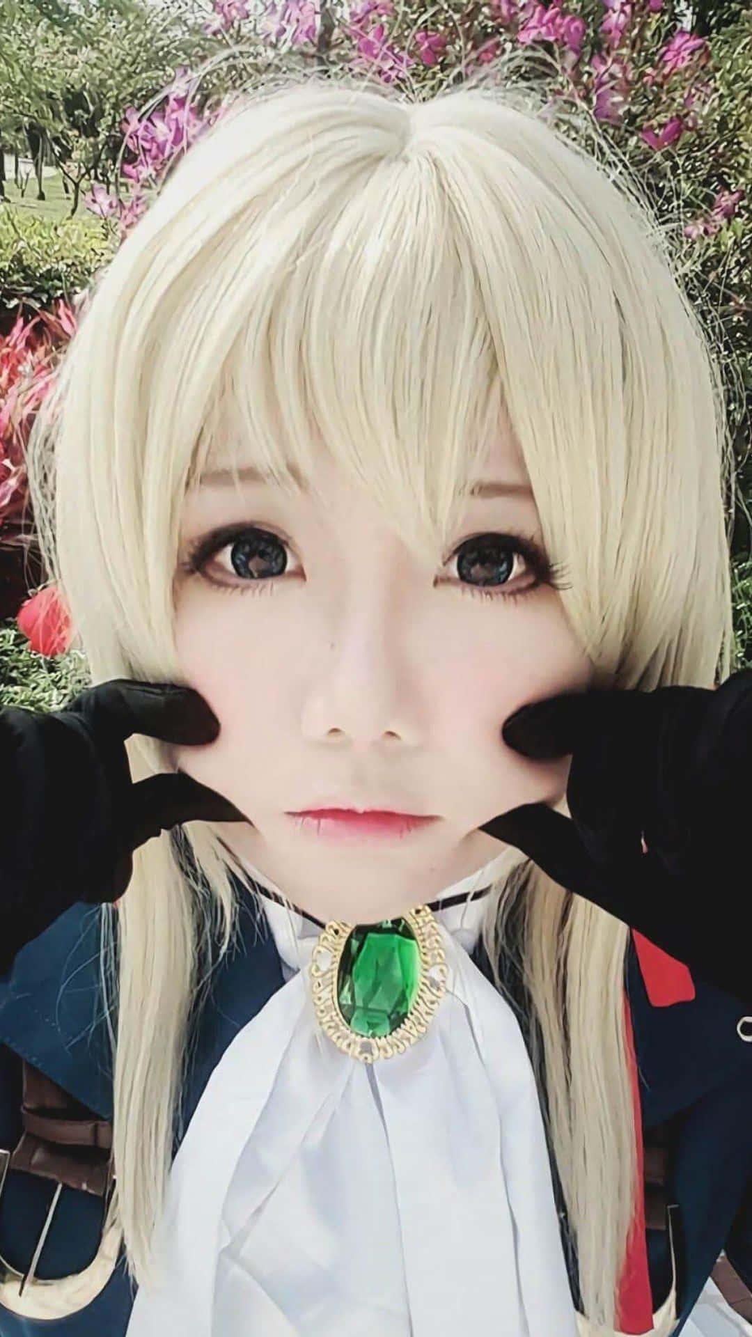 Cosplay Anime Kawaii 桌布 壁紙 — 一位擁有鉑金色頭髮和綠色寶石胸針的角色扮演者，戴著手套的雙手框著臉龐，在粉紅色花叢前擺姿勢。