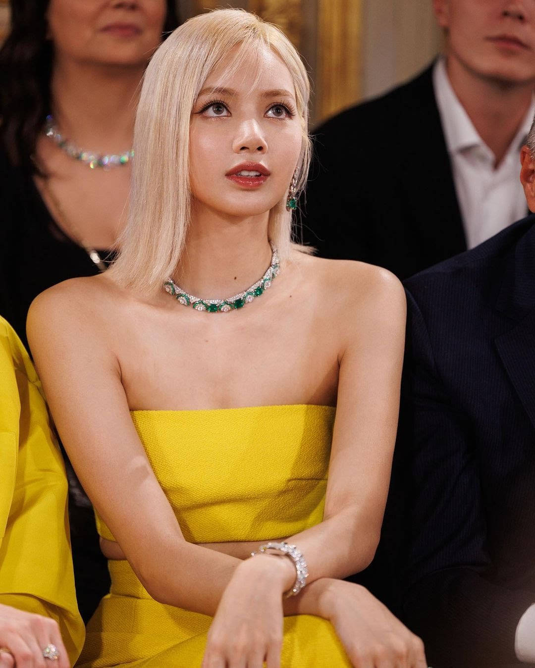 Beautiful Woman 桌布 壁紙 — 一位擁有鉑金色頭髮的女性穿著鮮黃色無肩帶連衣裙和翡翠珠寶，坐在優雅的活動中。