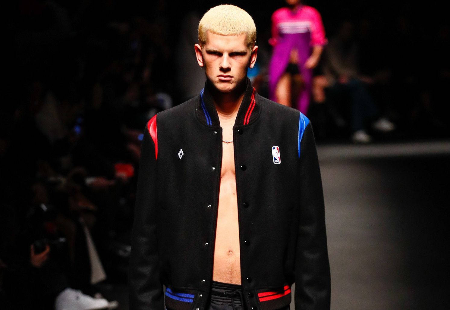 Fashion Model 桌布 壁紙 — 一位擁有鉑金色頭髮的模特兒自信地走在黑暗的伸展台上，身穿一件帶有紅藍色點綴和NBA標誌的黑色開襟Varsity外套。