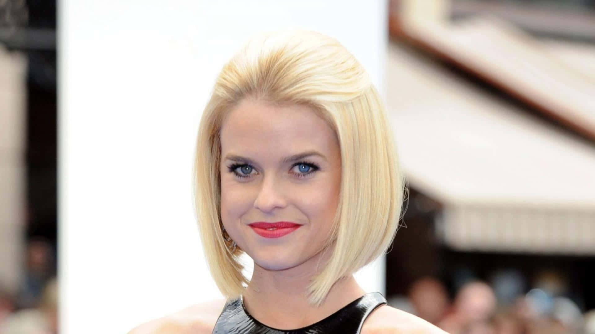 alice eve 桌布 壁紙 — 一位擁有鉑金色鮑伯頭和鮮豔紅唇的女性，在模糊的城市背景下溫柔微笑。