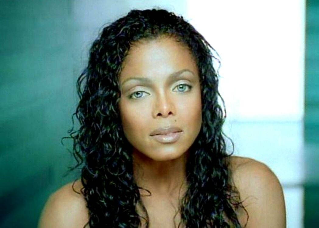 janet jackson 桌布 壁紙 — 一位擁有長捲曲黑髮和引人注目的綠色眼睛的女性，在柔和的青綠色和白色模糊背景前直視鏡頭。