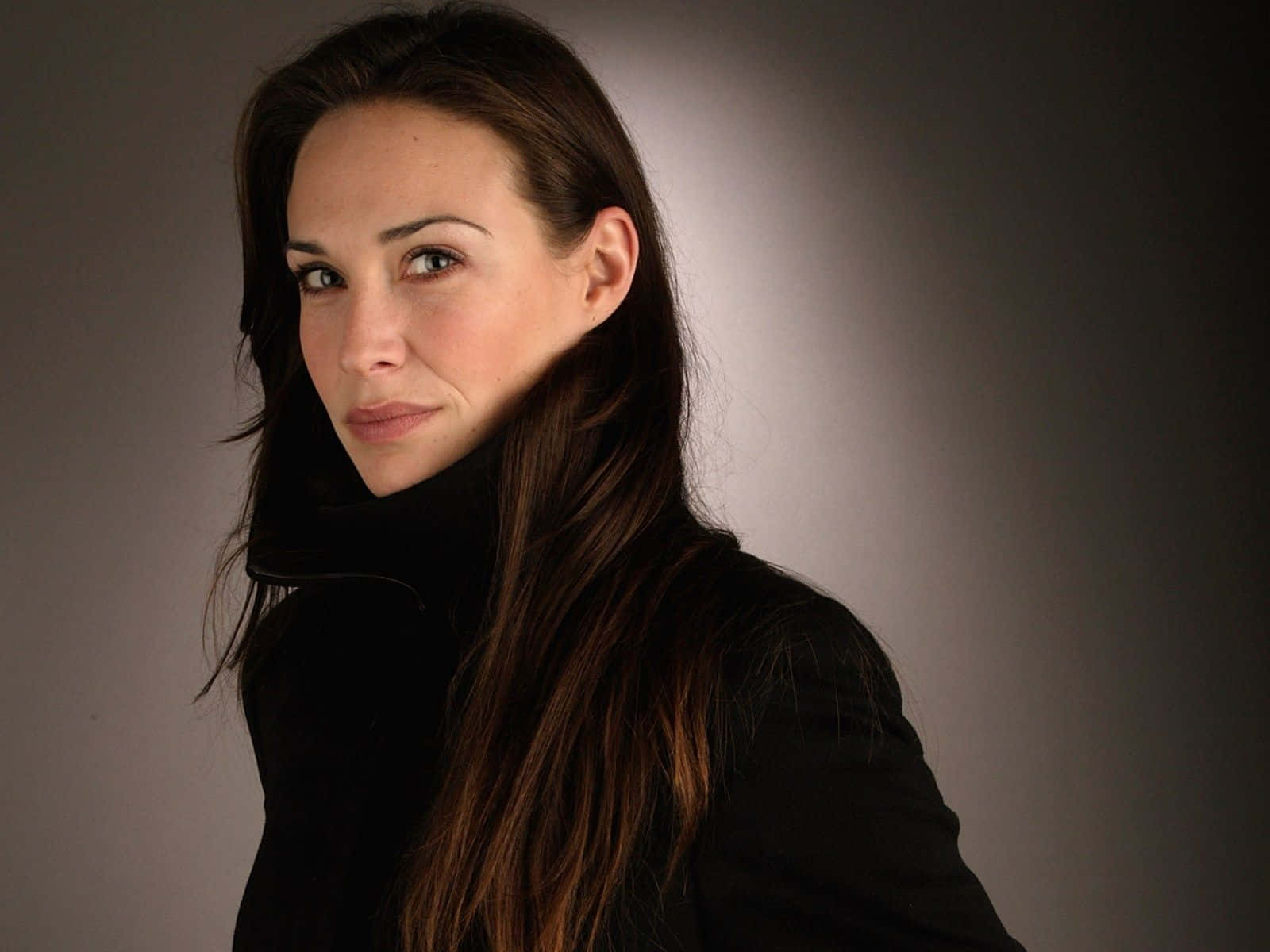 Claire Forlani 桌布 壁紙 — 一位擁有長棕髮、身穿黑色高領毛衣的女性，在柔和光線的灰色背景前直視鏡頭。