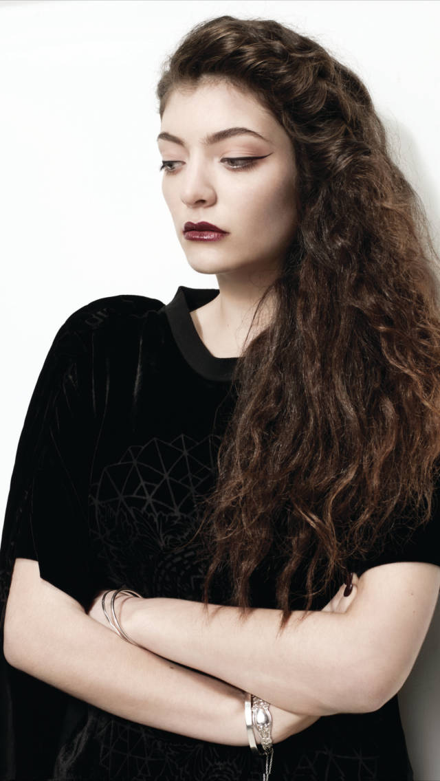Lorde 桌布 壁紙 — 一位擁有長波浪棕色頭髮和大膽紅唇的女性，站在純白色背景前，穿著帶有幾何刺繡的黑色天鵝絨上衣和銀色手鐲。