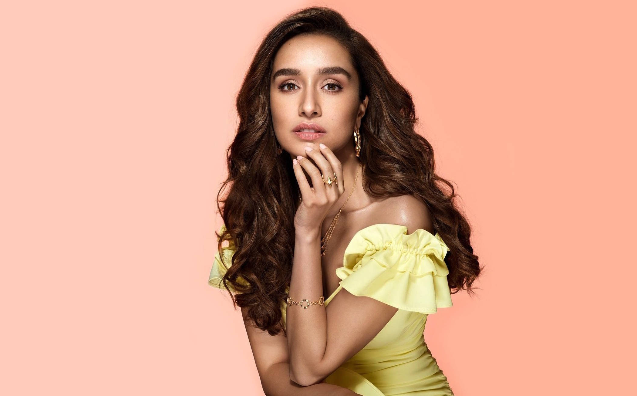Shraddha Kapoor 桌布 壁紙 — 一位擁有長波浪棕色頭髮的女性，在柔和的桃色背景前穿著黃色露肩連衣裙。