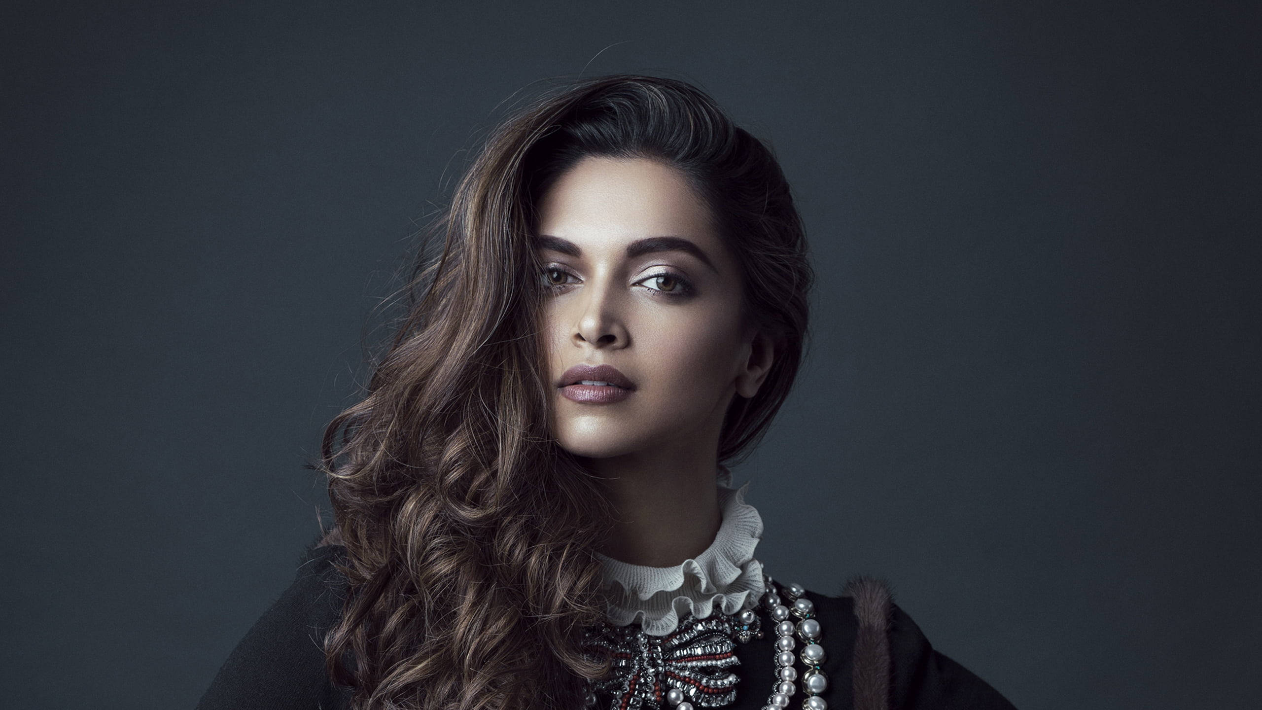 Deepika 桌布 壁紙 — 一位擁有長波浪棕色頭髮的女性在深灰色工作室背景前直視鏡頭。