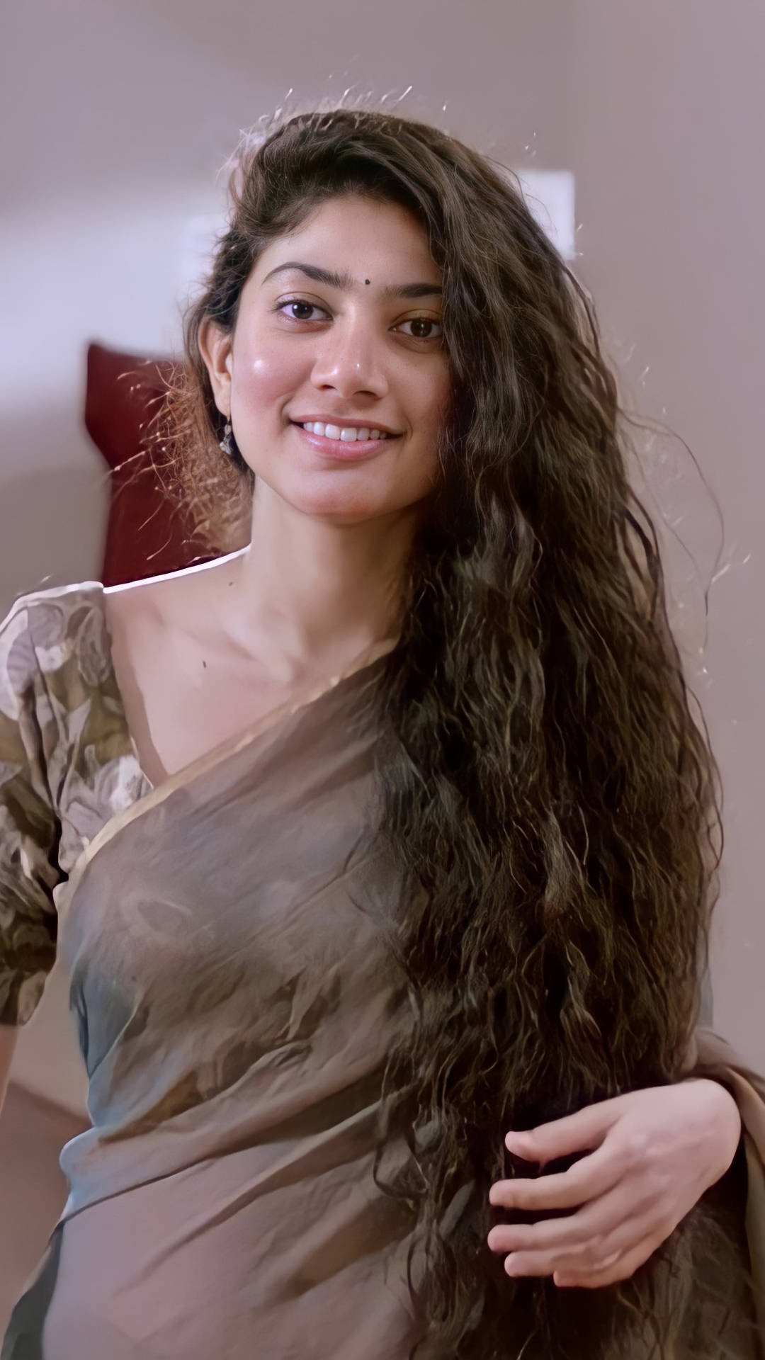 Sai Pallavi 桌布 壁紙 — 一位擁有長波浪深色頭髮的女性，在室內穿著透視棕色紗麗，溫暖地微笑。