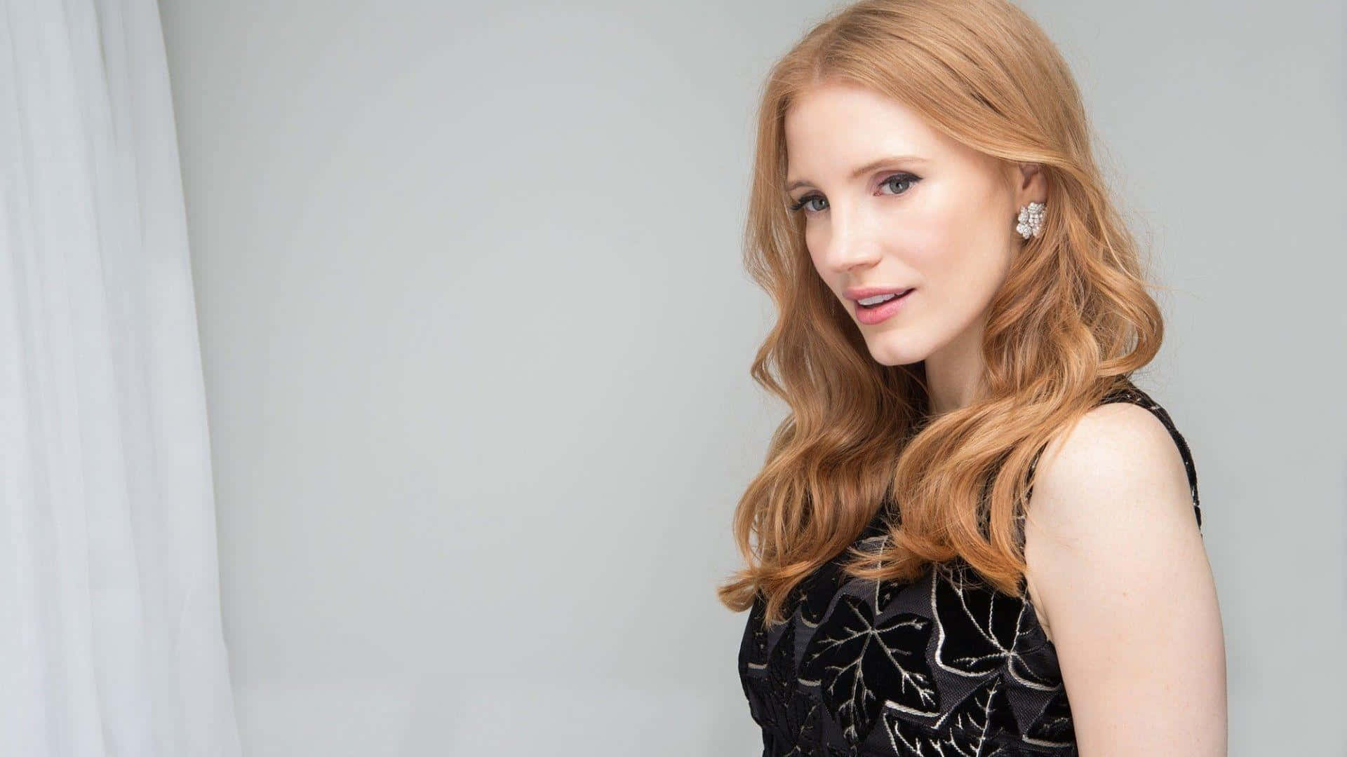 jessica chastain 桌布 壁紙 — 一位擁有長波浪紅髮的女性，穿著帶有銀色葉子刺繡的黑色連衣裙和閃亮耳環，背景是純灰色。