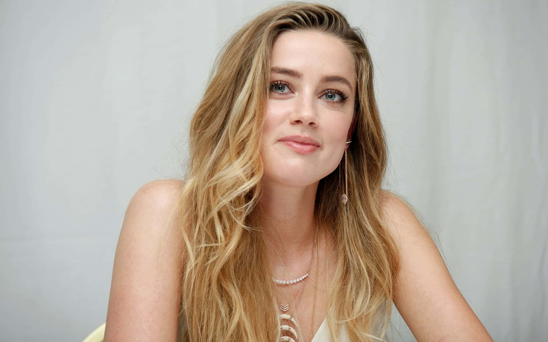 Amber Heard 桌布 壁紙 — 一位擁有長波浪金髮和藍眼睛的女性，在純淺灰色背景前溫柔微笑。