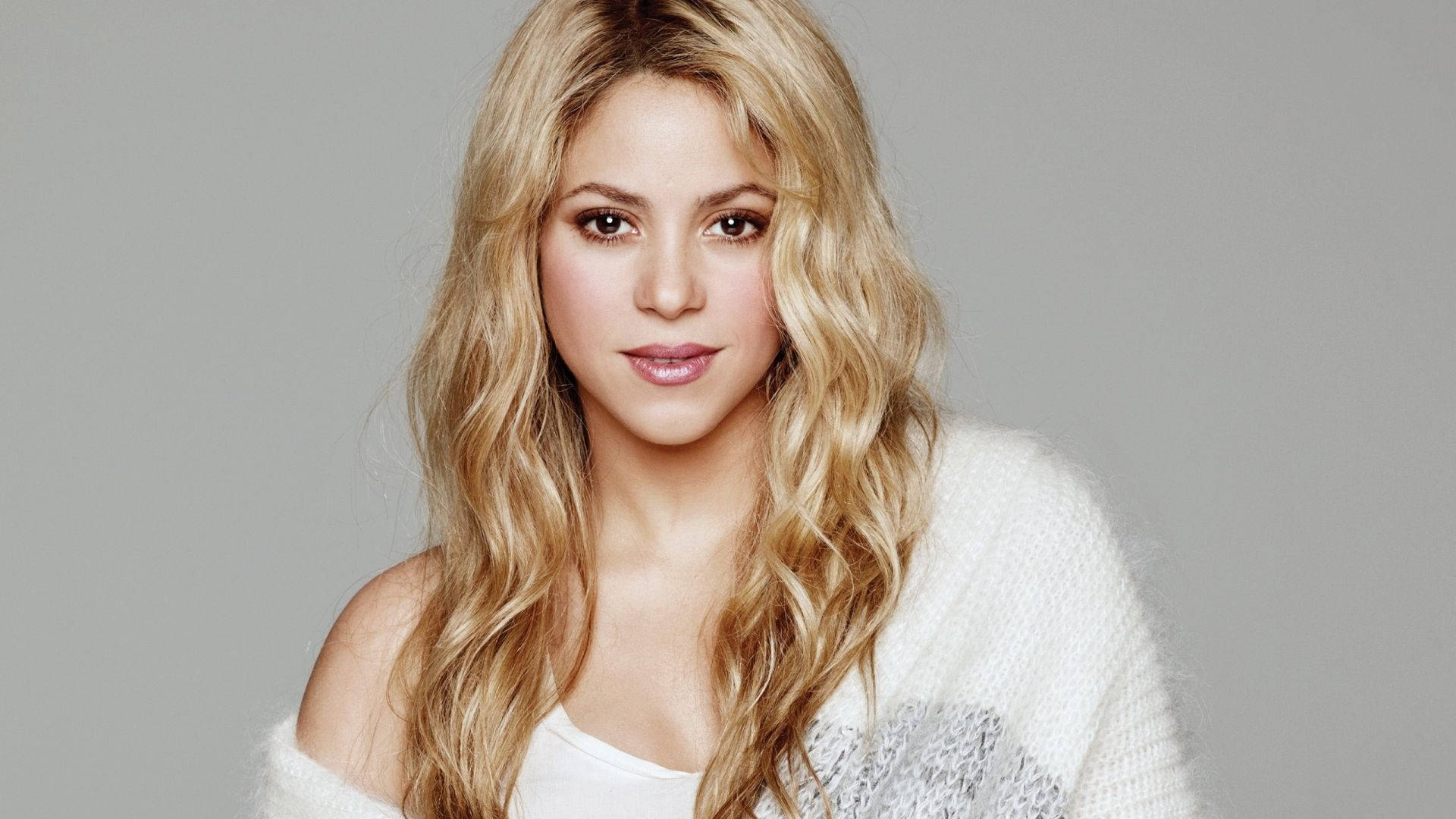 Shakira 桌布 壁紙 — 一位擁有長波浪金髮的女性，在純灰色背景前穿著白色毛絨毛衣。
