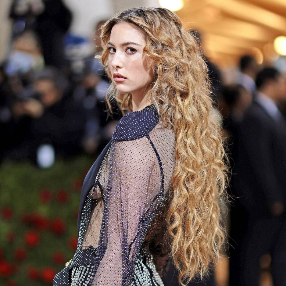 Curly Hair 桌布 壁紙 — 一位擁有長波浪金髮的女性，在背景模糊的攝影師的華麗活動中，穿著閃閃發光的透視黑色連衣裙。