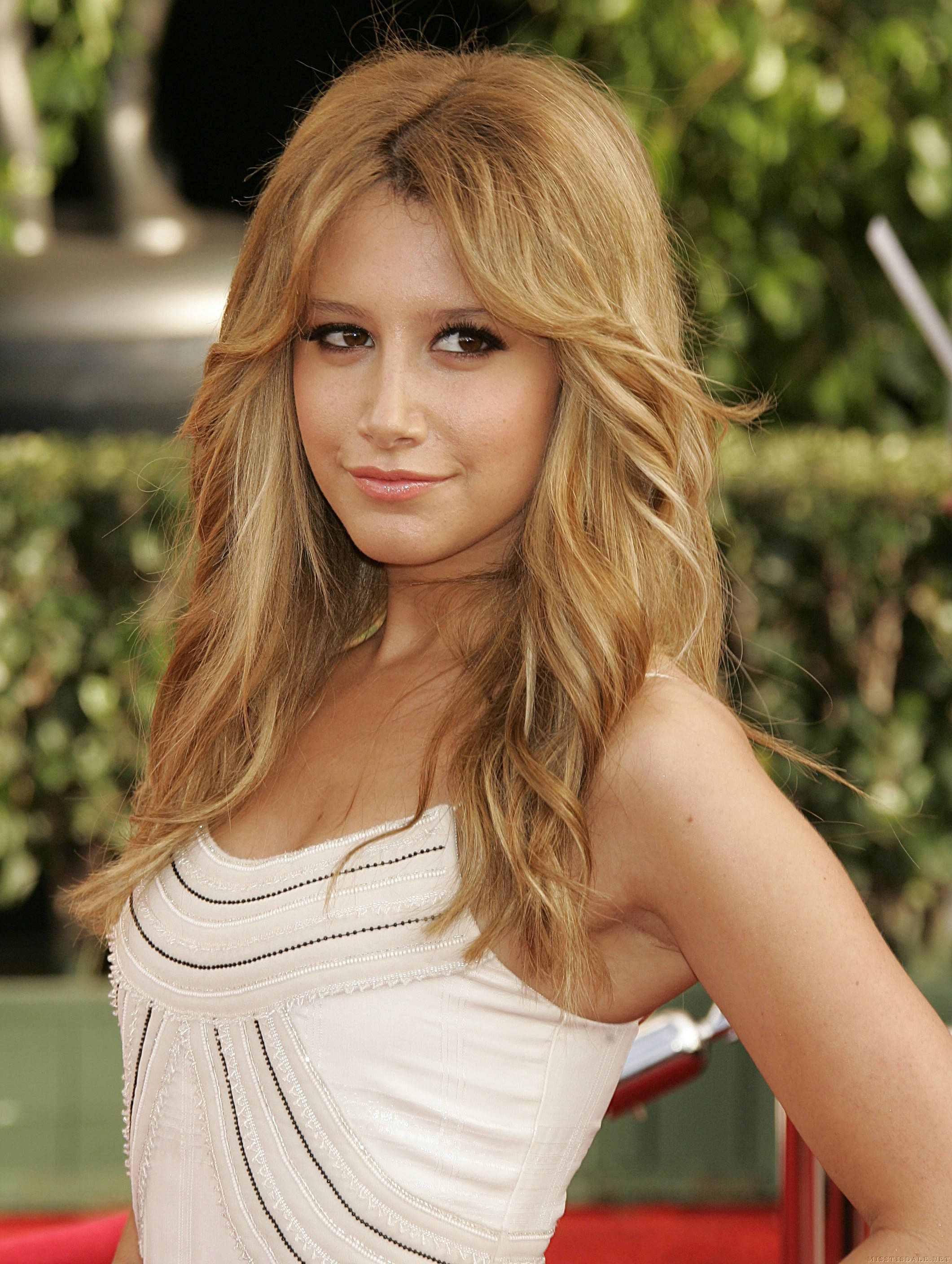 Ashley Tisdale 桌布 壁紙 — 一位擁有長波浪金髮的女性，穿著白色珠飾連衣裙，在綠色樹籬背景的紅地毯上溫柔微笑。