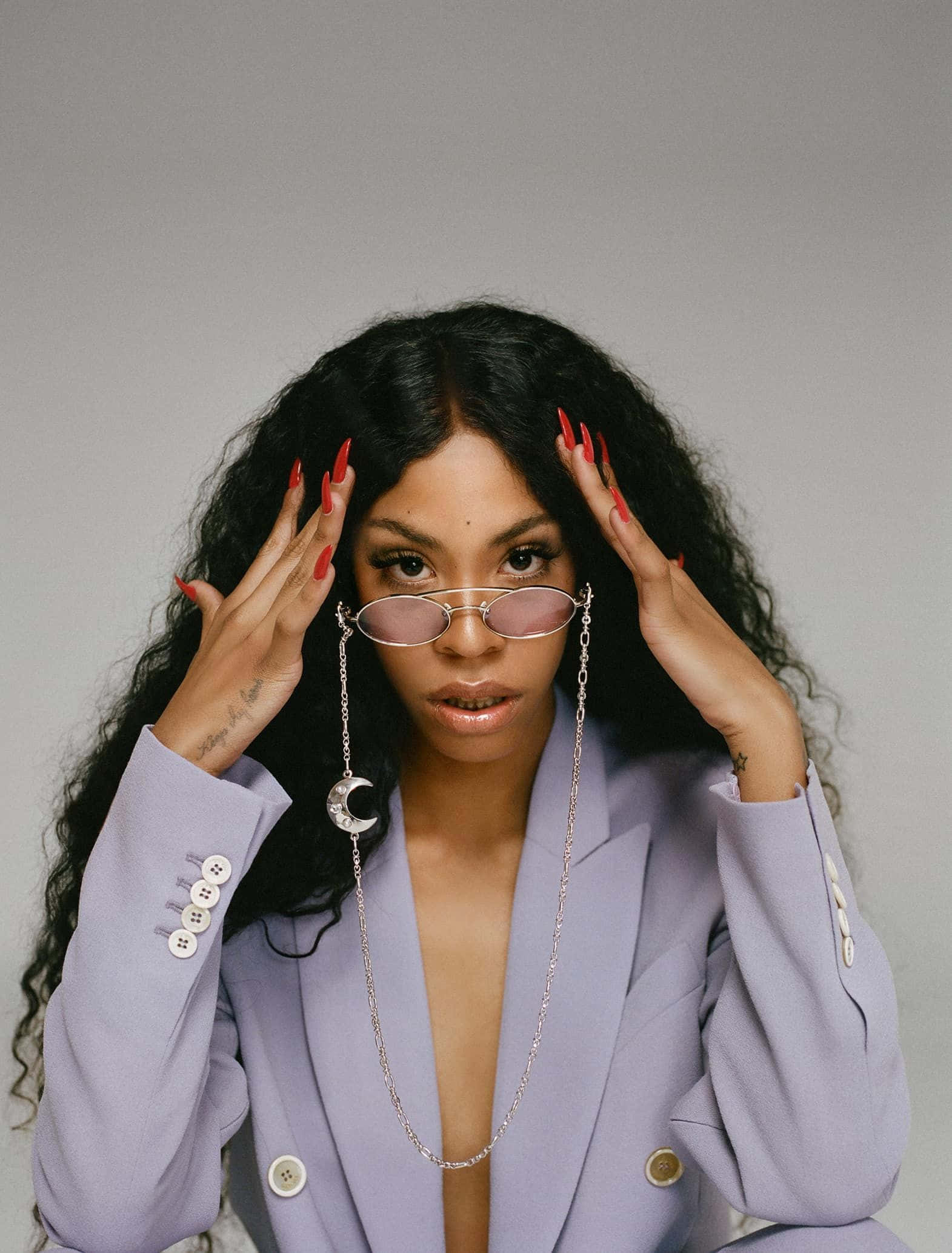 Rico Nasty 桌布 壁紙 — 一位擁有長深色捲髮和紅色指甲的女性，穿著薰衣草色西裝外套和粉色眼鏡，雙手抱著頭。