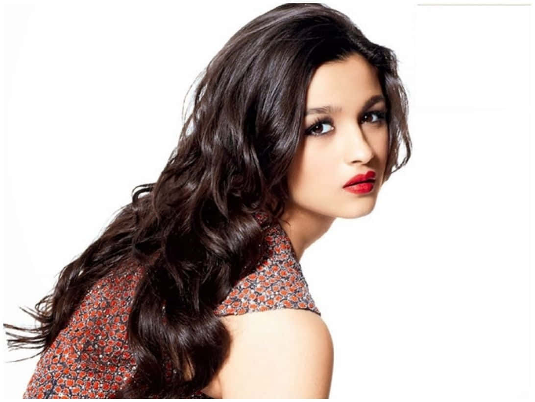 Alia Bhatt 桌布 壁紙 — 一位擁有長深色波浪頭髮和大膽紅唇的女性，在純白背景上回頭凝視。