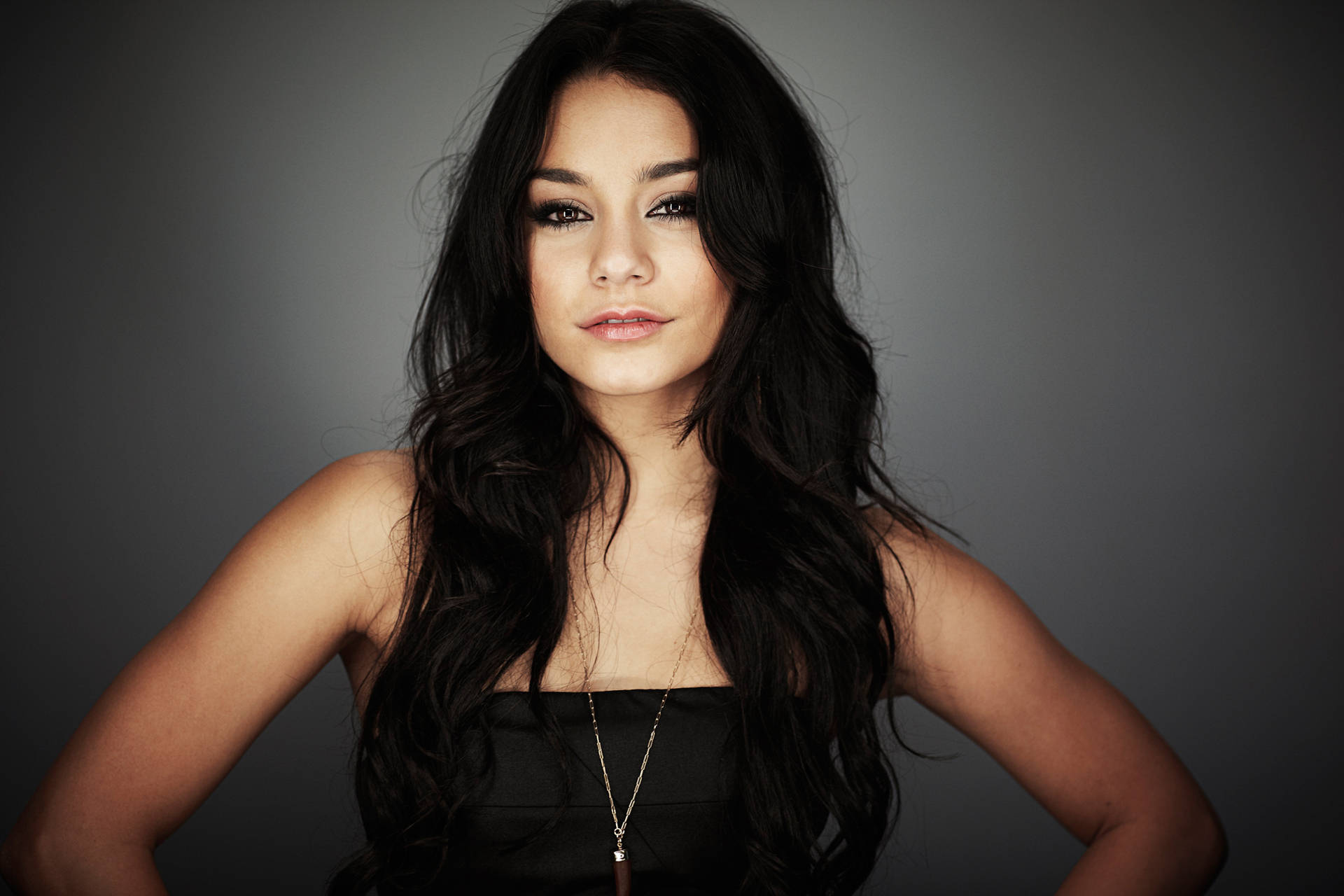 Vanessa Hudgens 桌布 壁紙 — 一位擁有長深色波浪髮和煙燻妝眼睛的女性，在柔和的灰色背景前自信地凝視鏡頭。