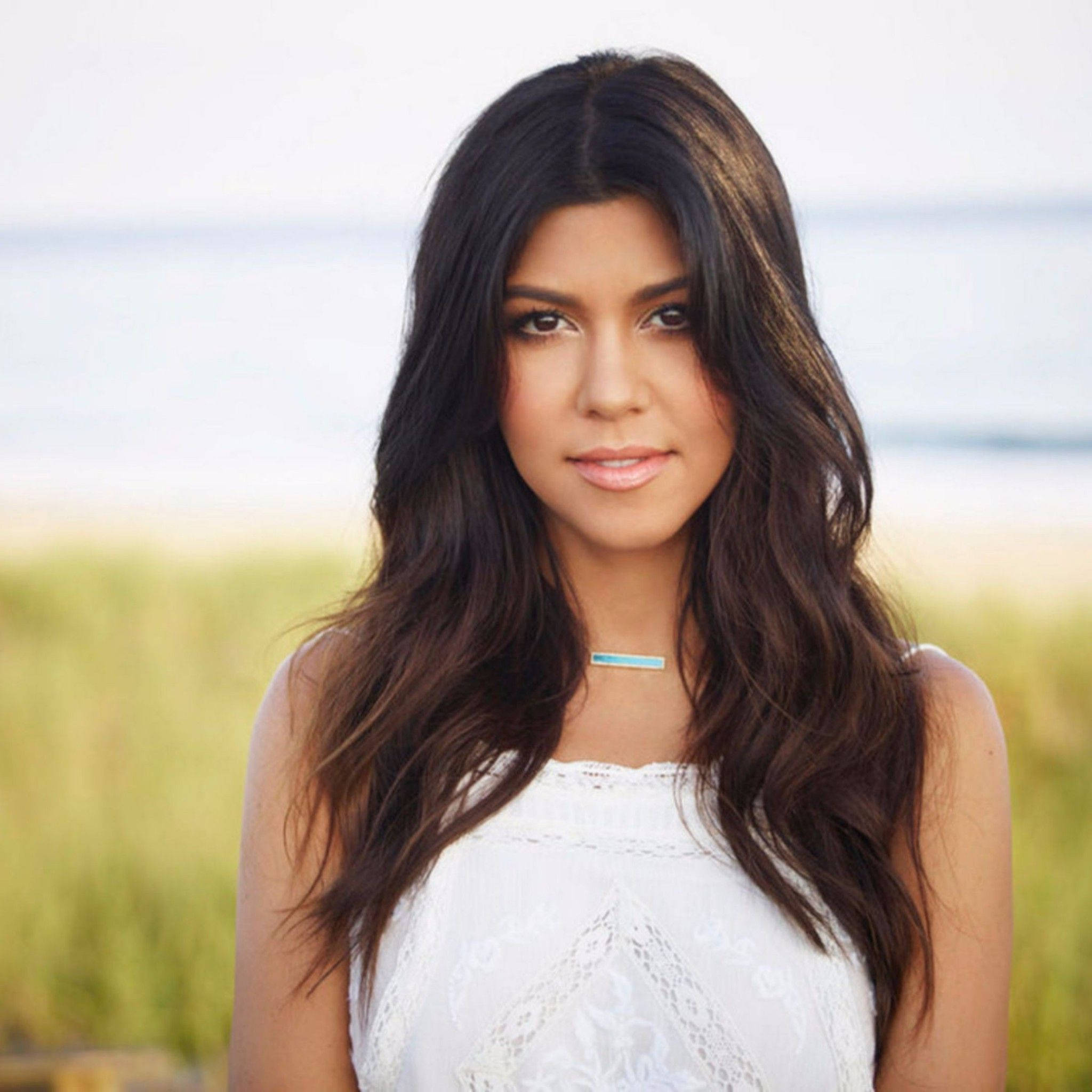 Kourtney 桌布 壁紙 — 一位擁有長深色波浪髮的女性，穿著白色蕾絲上衣和綠松石項鍊，背景是模糊的海灘。