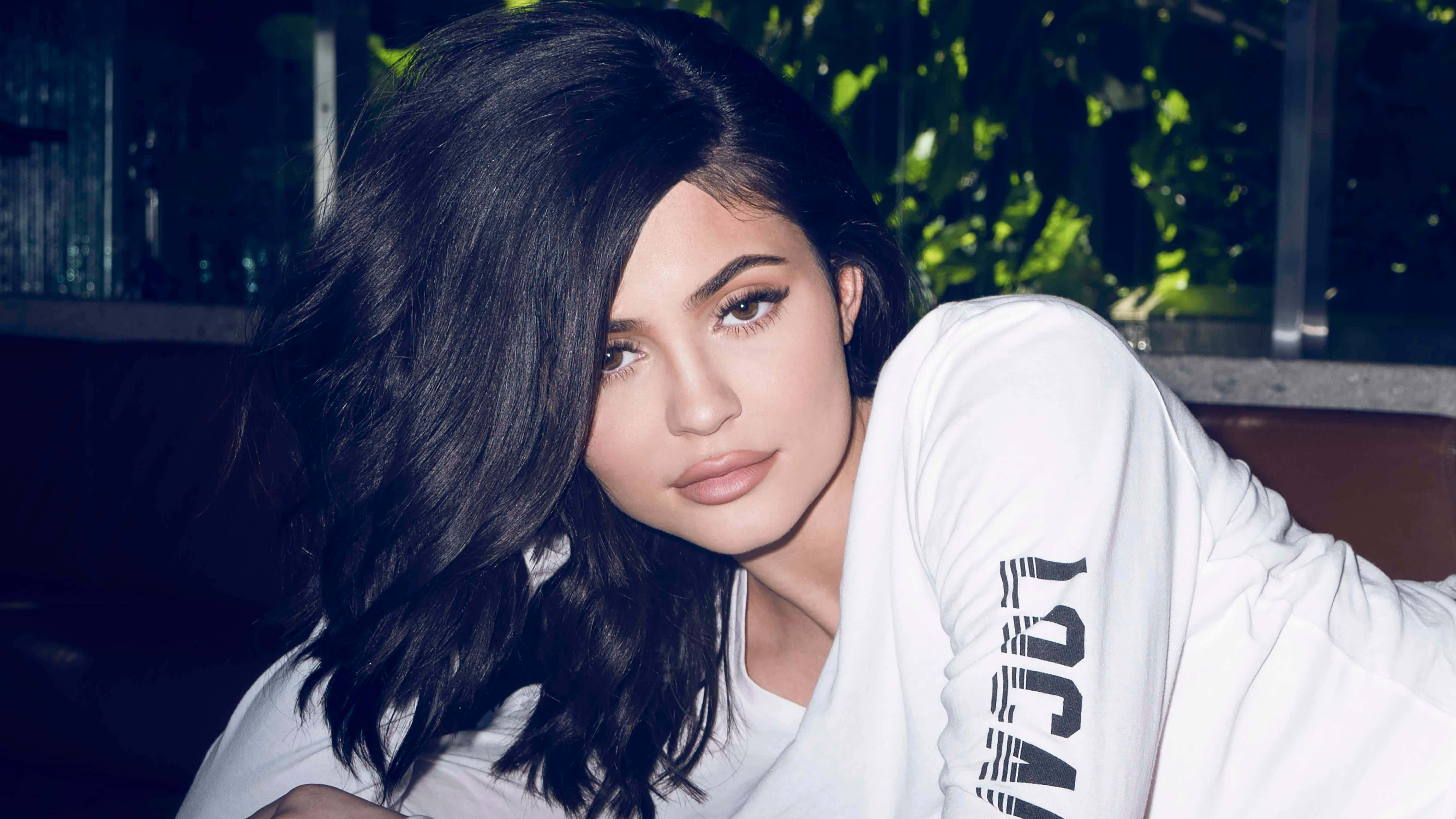 Kylie Jenner 4K 桌布 壁紙 — 一位擁有長深色波浪髮的女性向前傾，身穿帶有黑色文字的白色長袖襯衫。