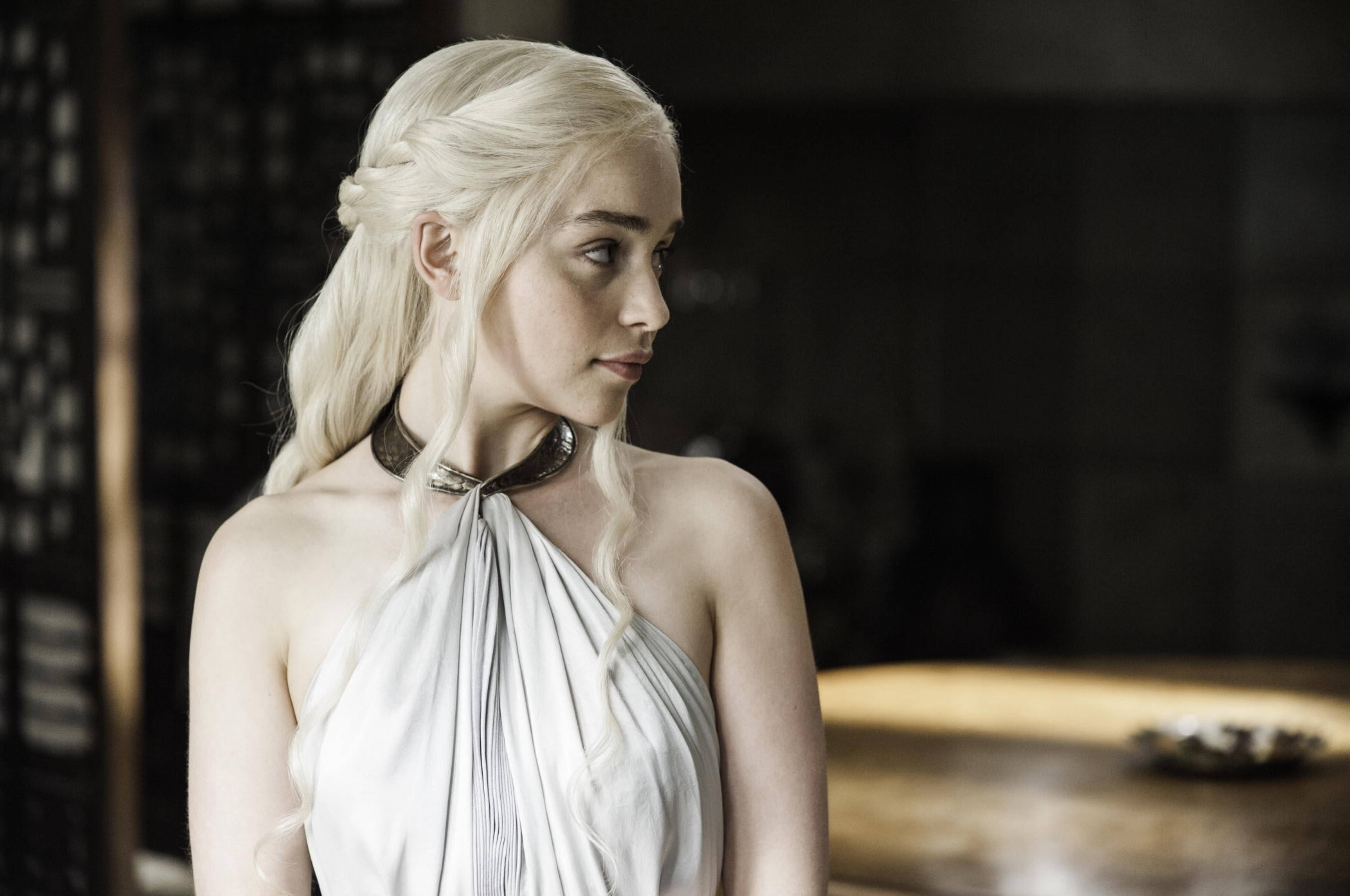daenerys 桌布 壁紙 — 一位擁有長白金髮的女性，身穿白色垂褶長袍和深色頸鏈，在昏暗的房間裡若有所思地側視。